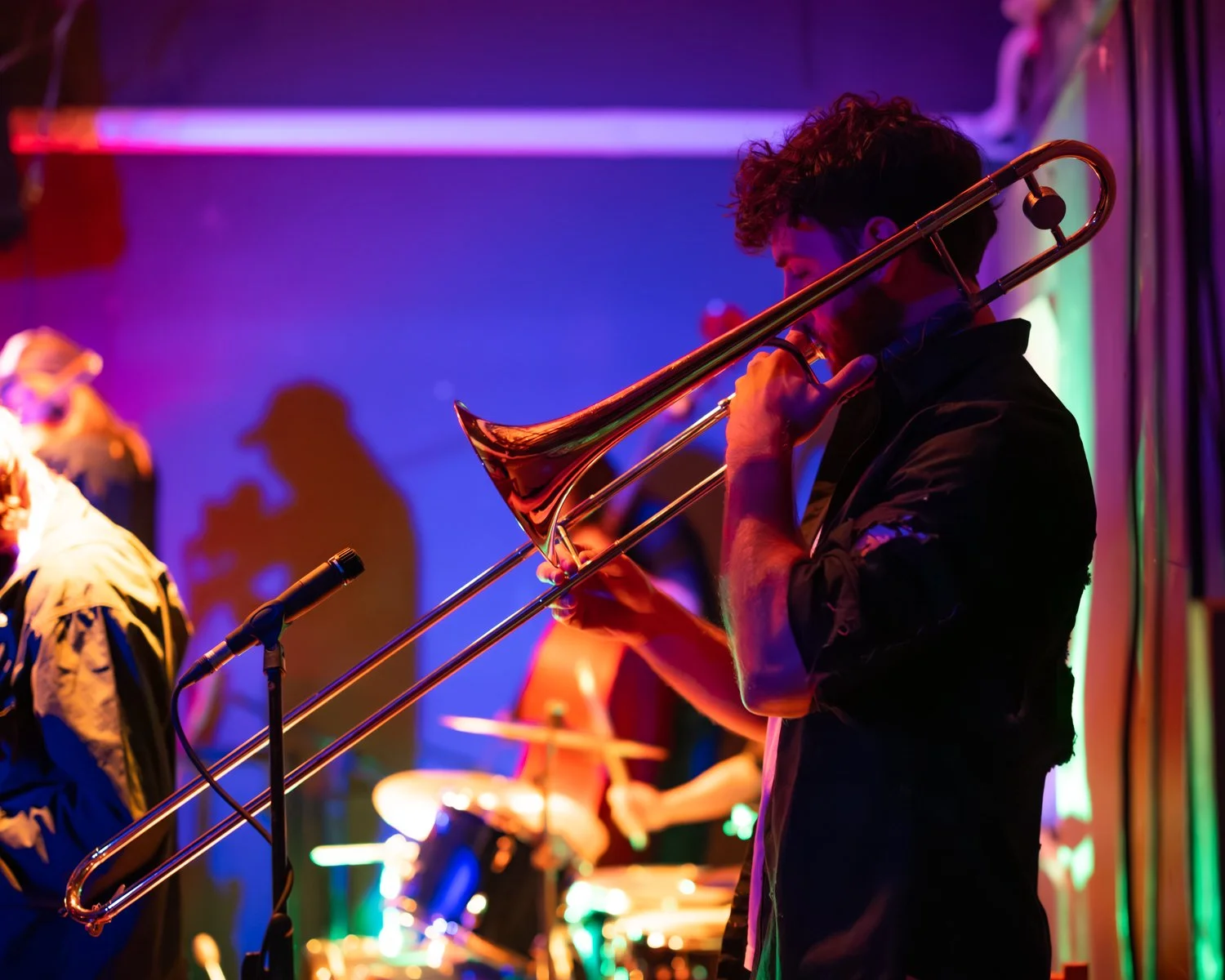A musician playing the trombone on stage with colorful lights and other band members in the background.