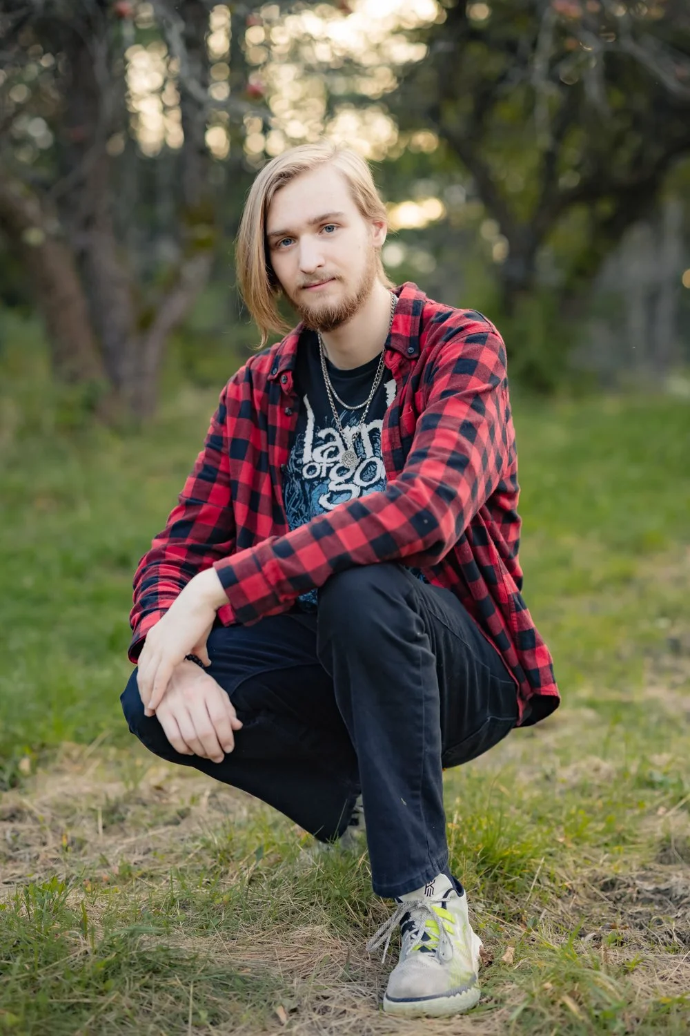A young man with long blonde hair and a beard crouching on grass in a park, wearing a red and black checkered shirt, a black graphic t-shirt, black pants, and white sneakers, with trees and sunlight in the background.