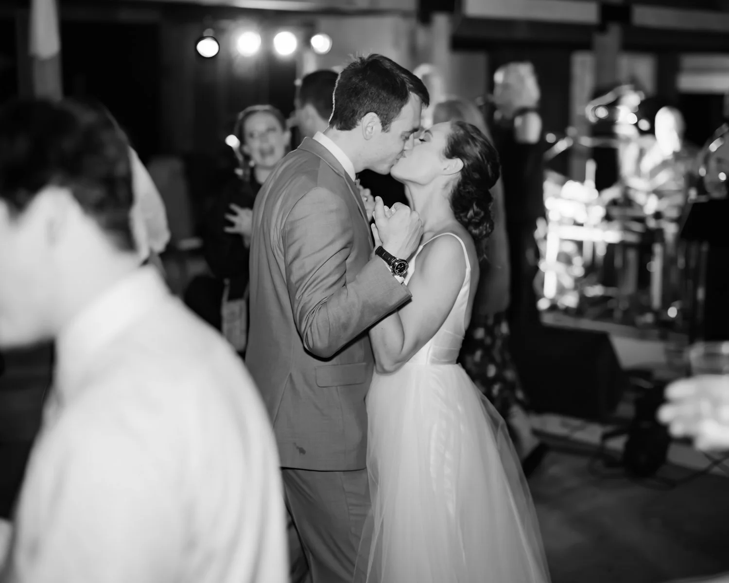 A black-and-white photo of a bride and groom sharing a kiss during their wedding reception, surrounded by guests and musicians.