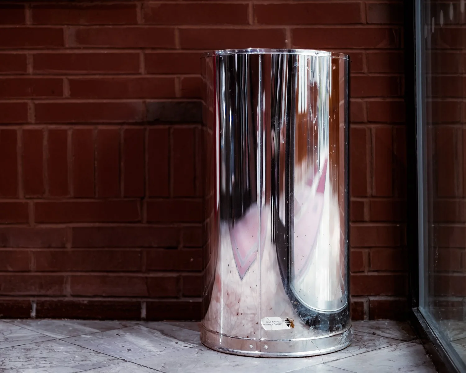 A shiny, reflective, metal trash can placed on a tiled floor in front of a red brick wall.