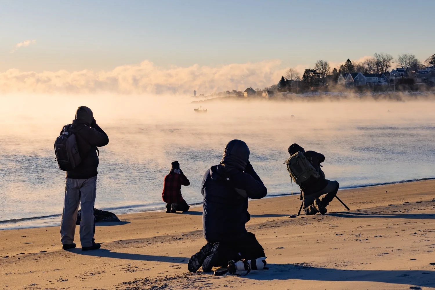 Four photographers on a sandy beach capturing a sunrise or sunset over misty water with houses on the distant shoreline in the background.