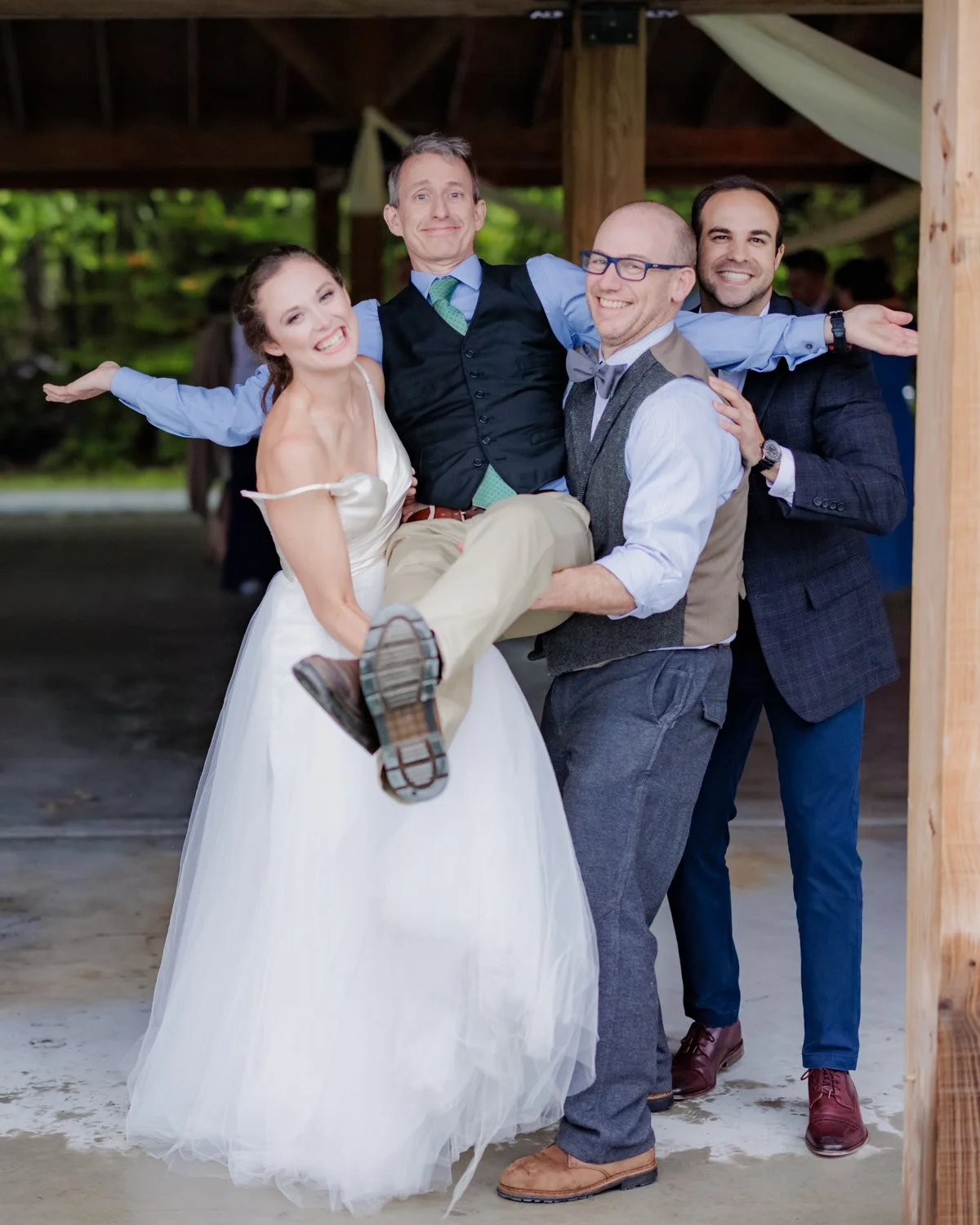 Four people at a wedding: a woman in a white wedding dress, and three men, one in a vest and glasses, all smiling and holding each other inside a wooden structure.
