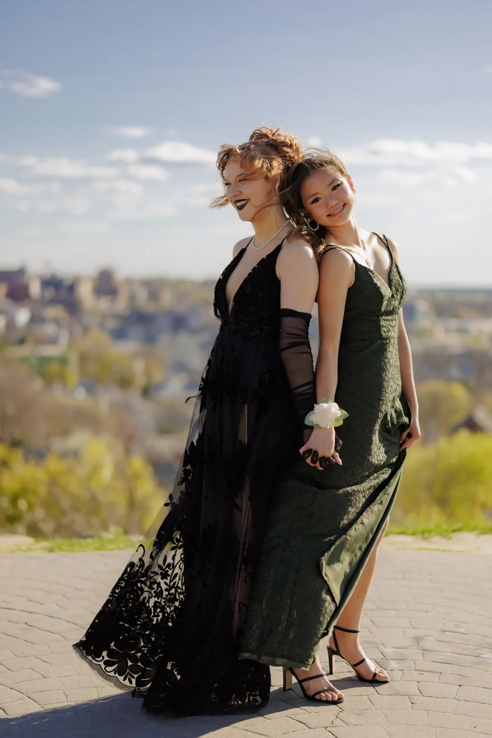 Two women in formal dresses standing outdoors on a sunny day, smiling and holding hands, with a cityscape and trees in the background.