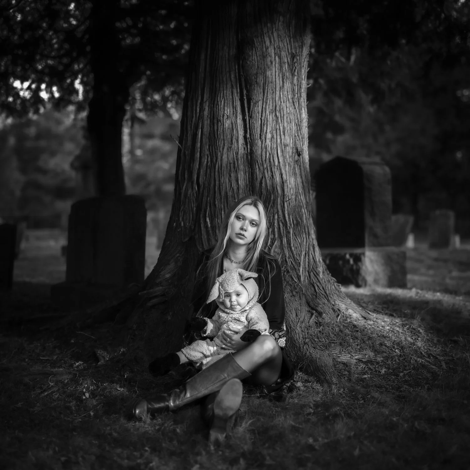 A young woman and a baby sitting on the ground in front of a large tree in a cemetery at dusk, in black and white.
