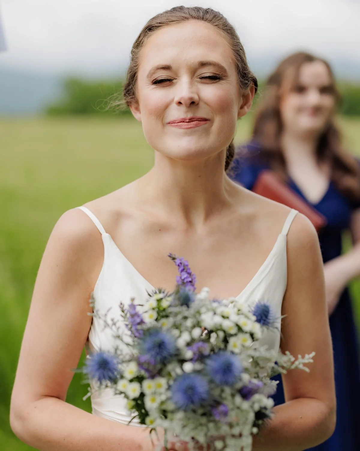 A woman in a white dress holding a bouquet of flowers on a grassy field with another woman smiling in the background.