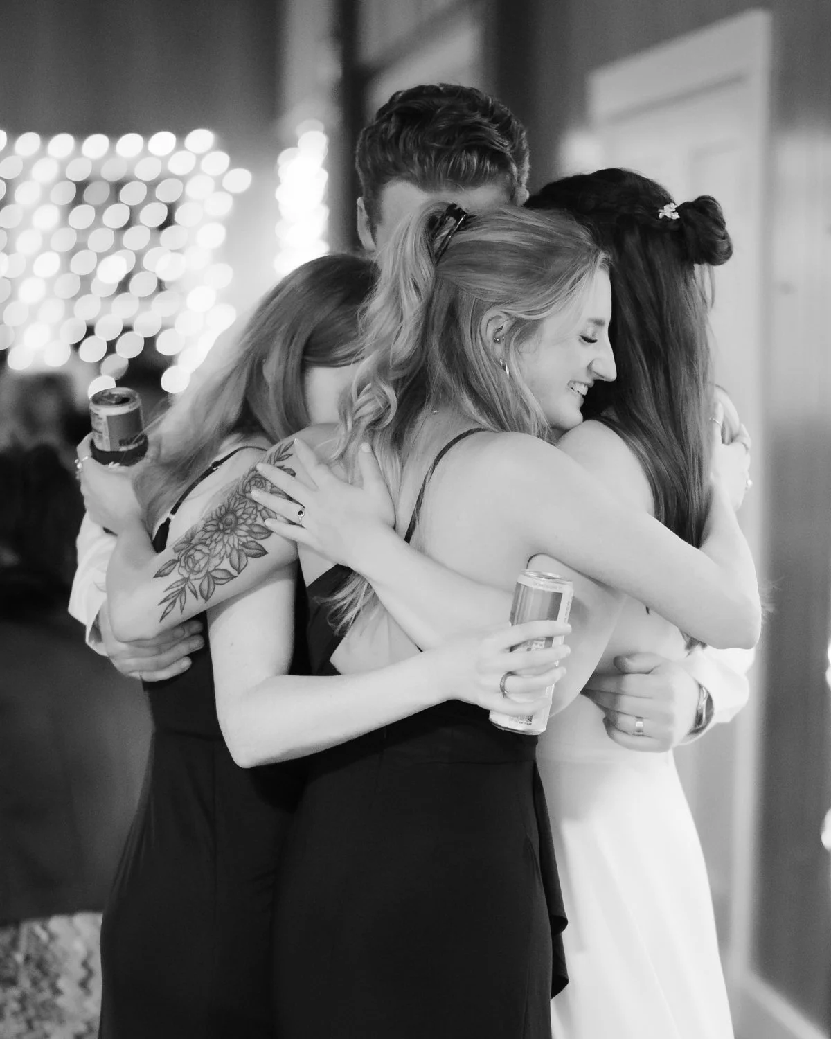 Four women hugging tightly at a celebration or gathering, some holding drinks, in a warmly lit indoor setting.