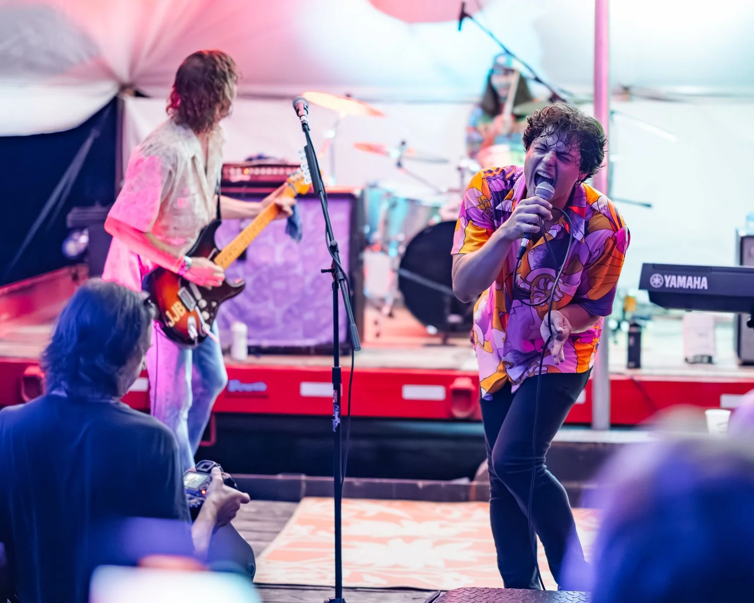 A band performs on stage under a tent, with a lead singer passionately singing into a microphone, a guitarist playing, and a drummer in the background, illuminated by colorful stage lights.
