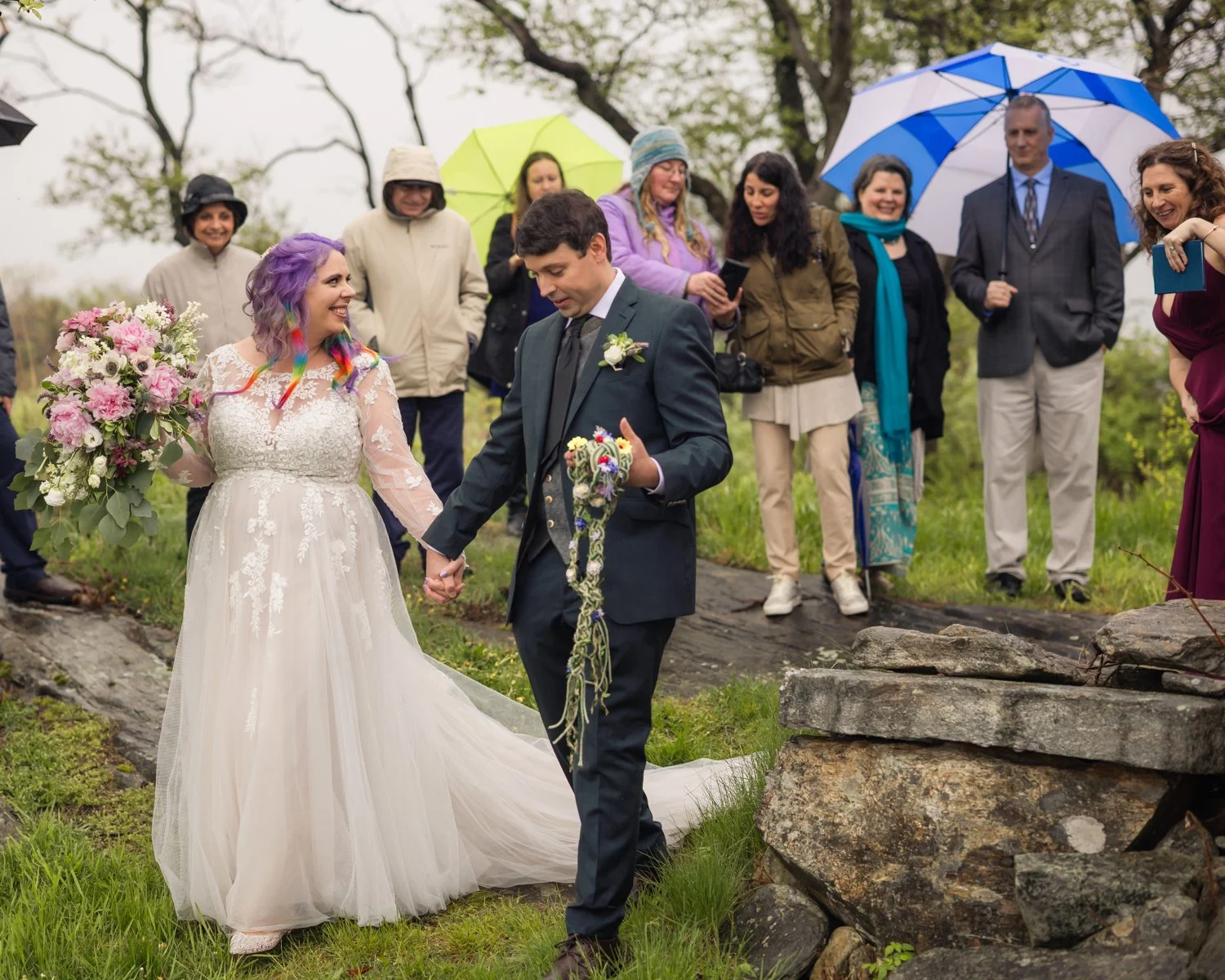 A bride and groom hold hands during their outdoor wedding ceremony, surrounded by friends and family holding umbrellas in an overcast setting with green grass and trees.