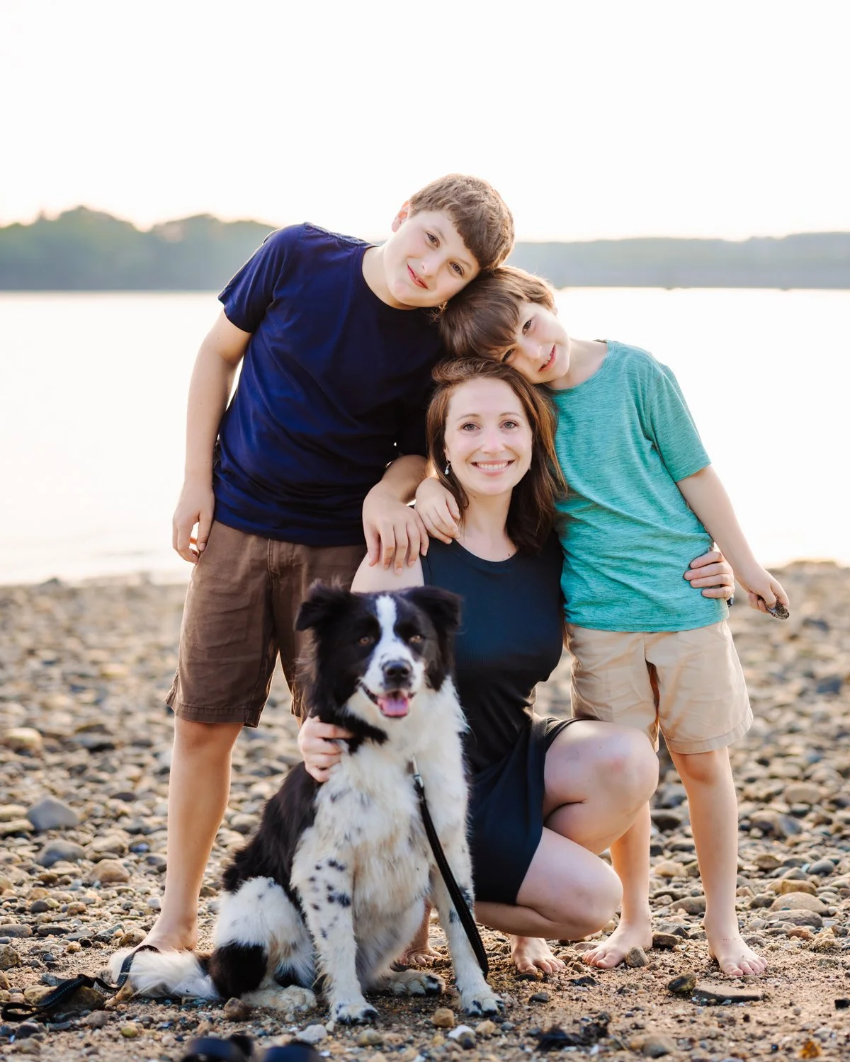 A smiling woman with two boys and a black and white dog on a rocky beach by a lake at sunset.