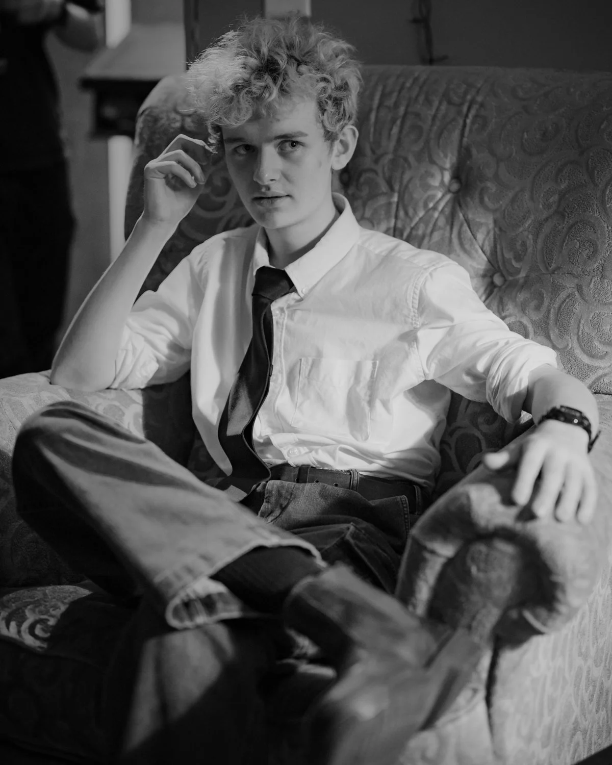 A young man with curly hair wearing a white shirt, loosened tie, and jeans, sitting on a sofa with his legs crossed. He is looking at the camera and touching his hair with one hand.