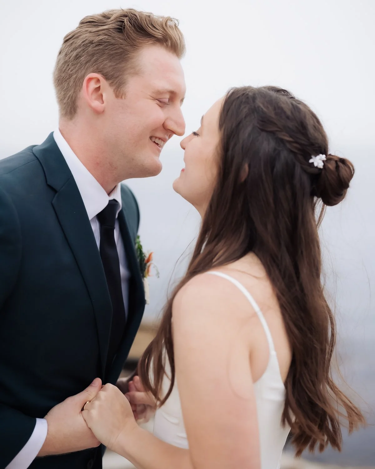 A couple in wedding attire facing each other outdoors, smiling, holding hands, with an overcast sky in the background.