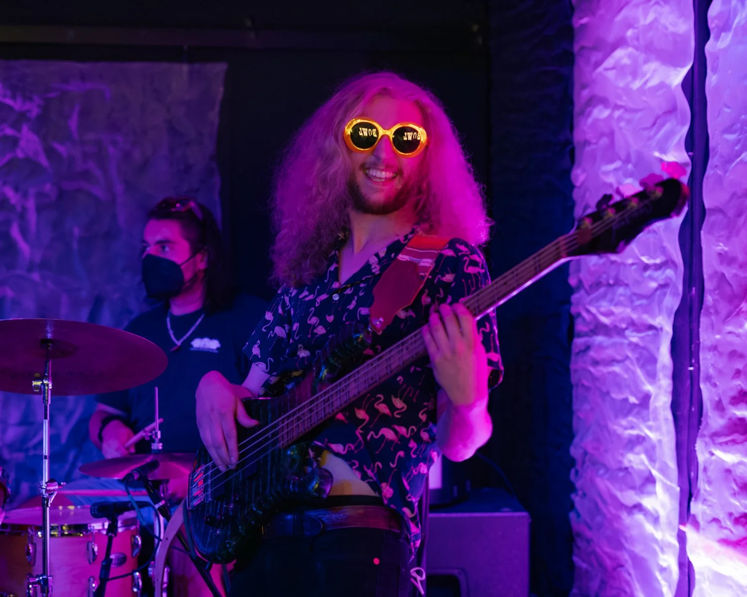 Person with curly hair wearing yellow sunglasses, playing an electric bass guitar during a live music performance in a colorful, dimly lit venue.