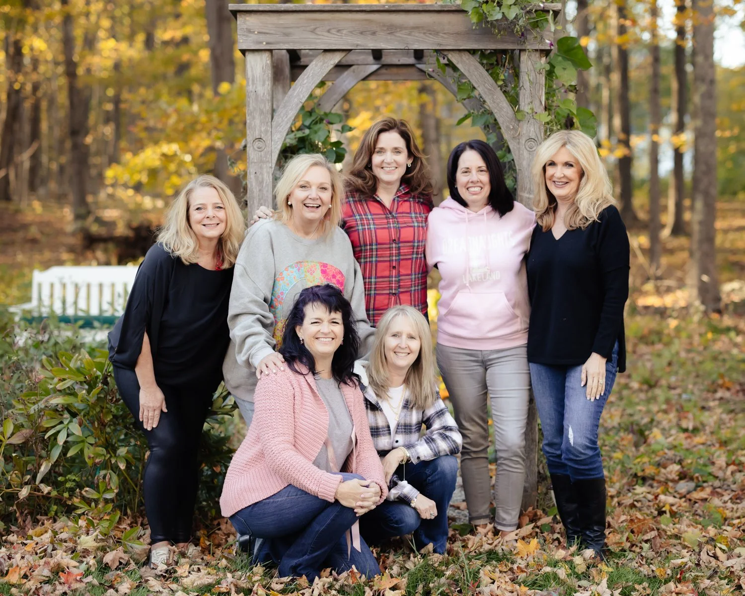 Group of eight women posing outdoors in a wooded area during fall, standing in front of a wooden arbor with vines. They are smiling and dressed casually, with autumn leaves on the ground.