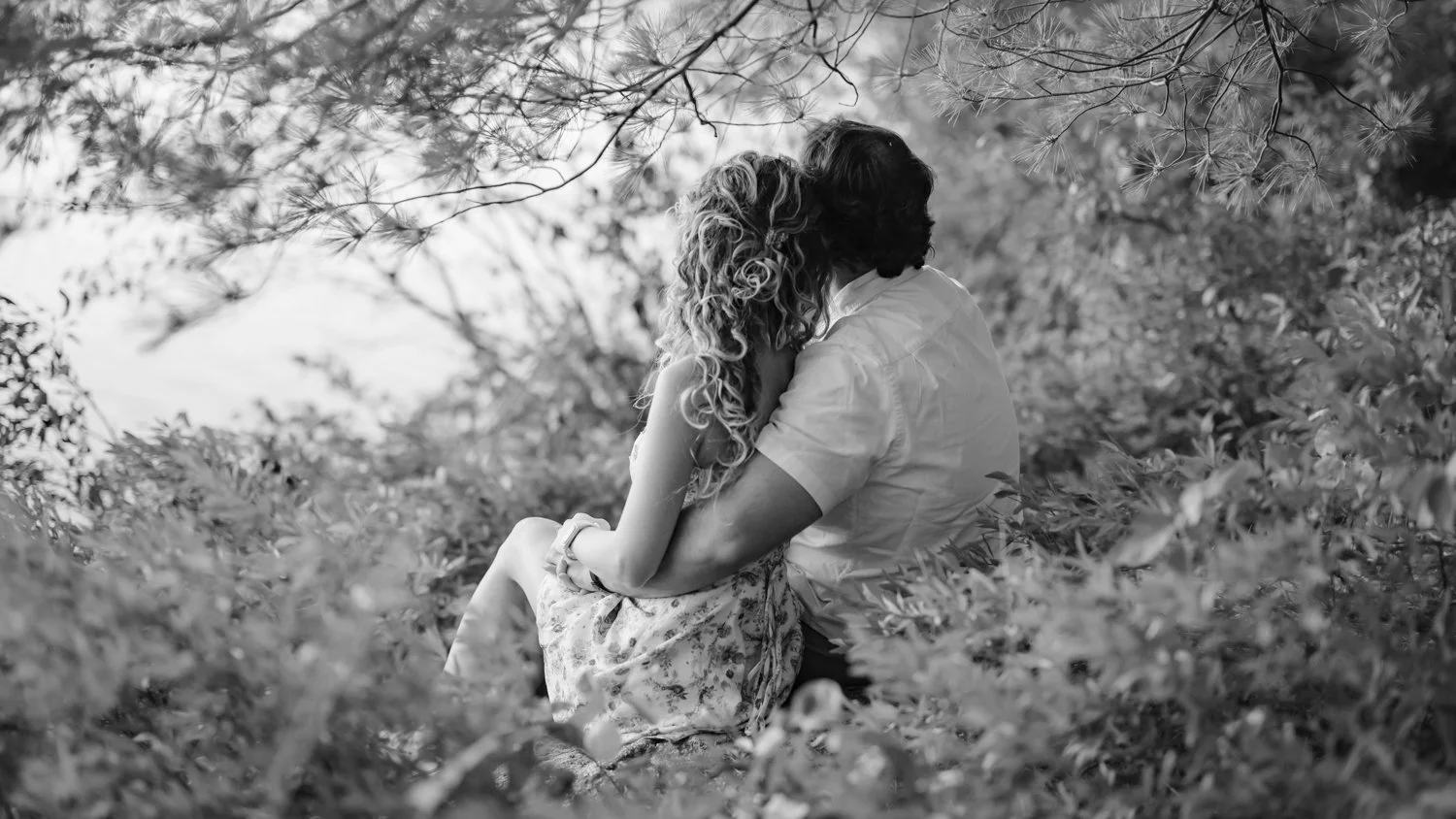 A black and white photo of a couple sitting close on the ground surrounded by trees and foliage, kissing or embracing amidst nature.
