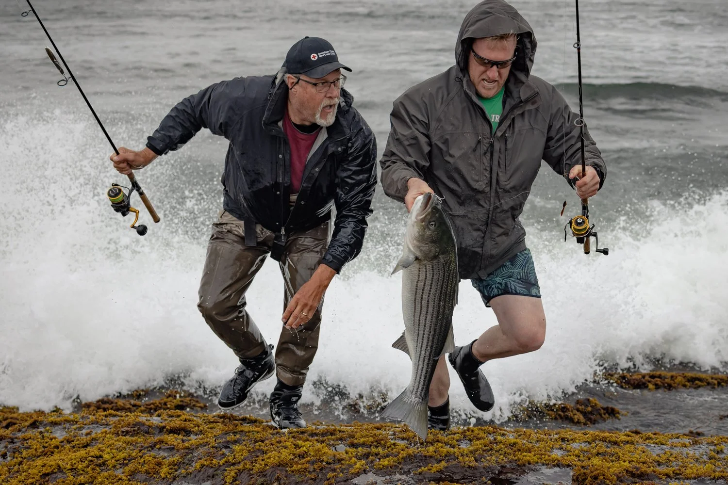 Two men in rain jackets and shorts catch a large fish along the shoreline, with waves crashing behind them.