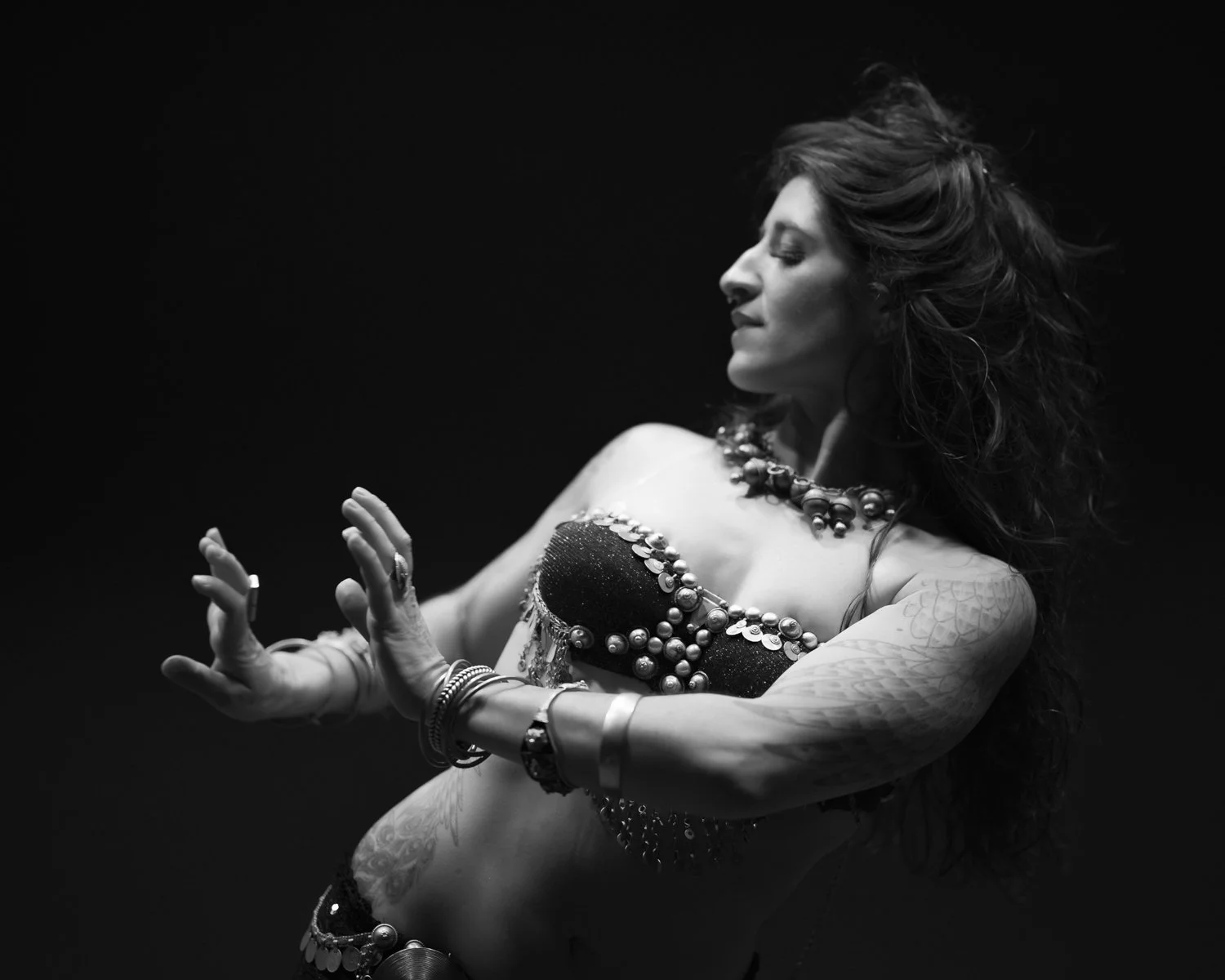 A woman with long curly hair in expressive dance pose, wearing a decorative top and accessories, against a dark background.