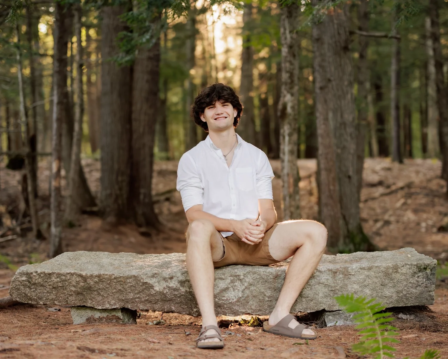 A young man sitting on a large rock in a forest, wearing a white shirt, brown shorts, and sandals, smiling at the camera with trees and sunlight in the background.