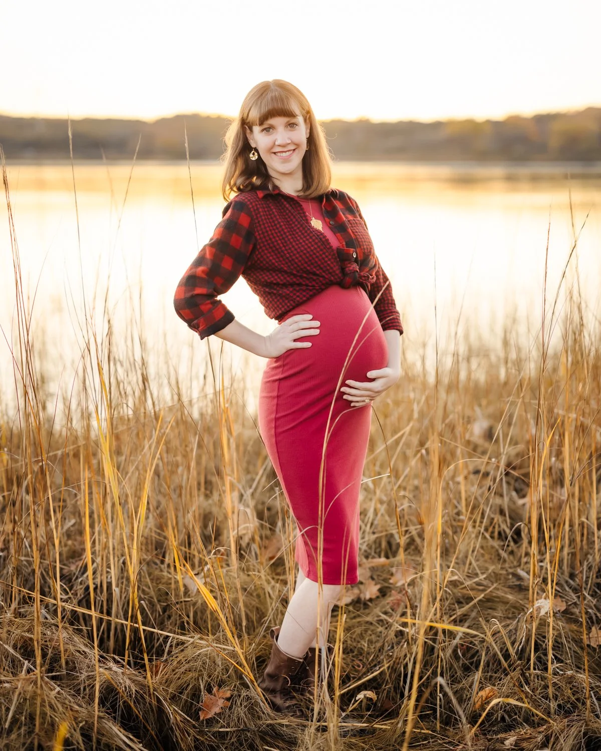 A pregnant woman with red hair standing in a field of tall, dry grass near a body of water during sunset, smiling and looking at the camera.