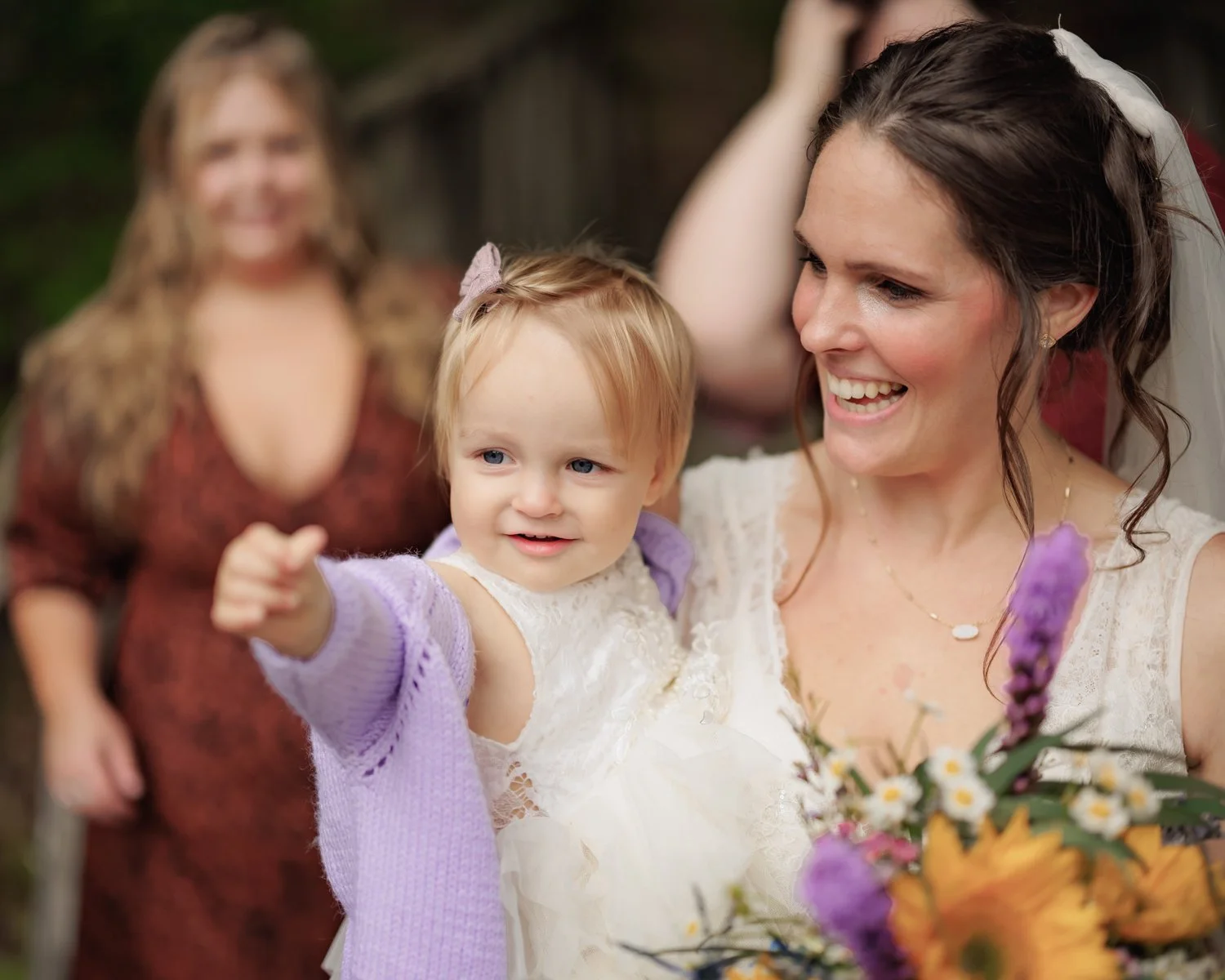 A bride smiling and holding a young girl, who is pointing. An older woman stands in the background, blurred. The bride wears a wedding dress and veil, and the young girl wears a white dress with a lavender cardigan. The bride holds a bouquet of flowe