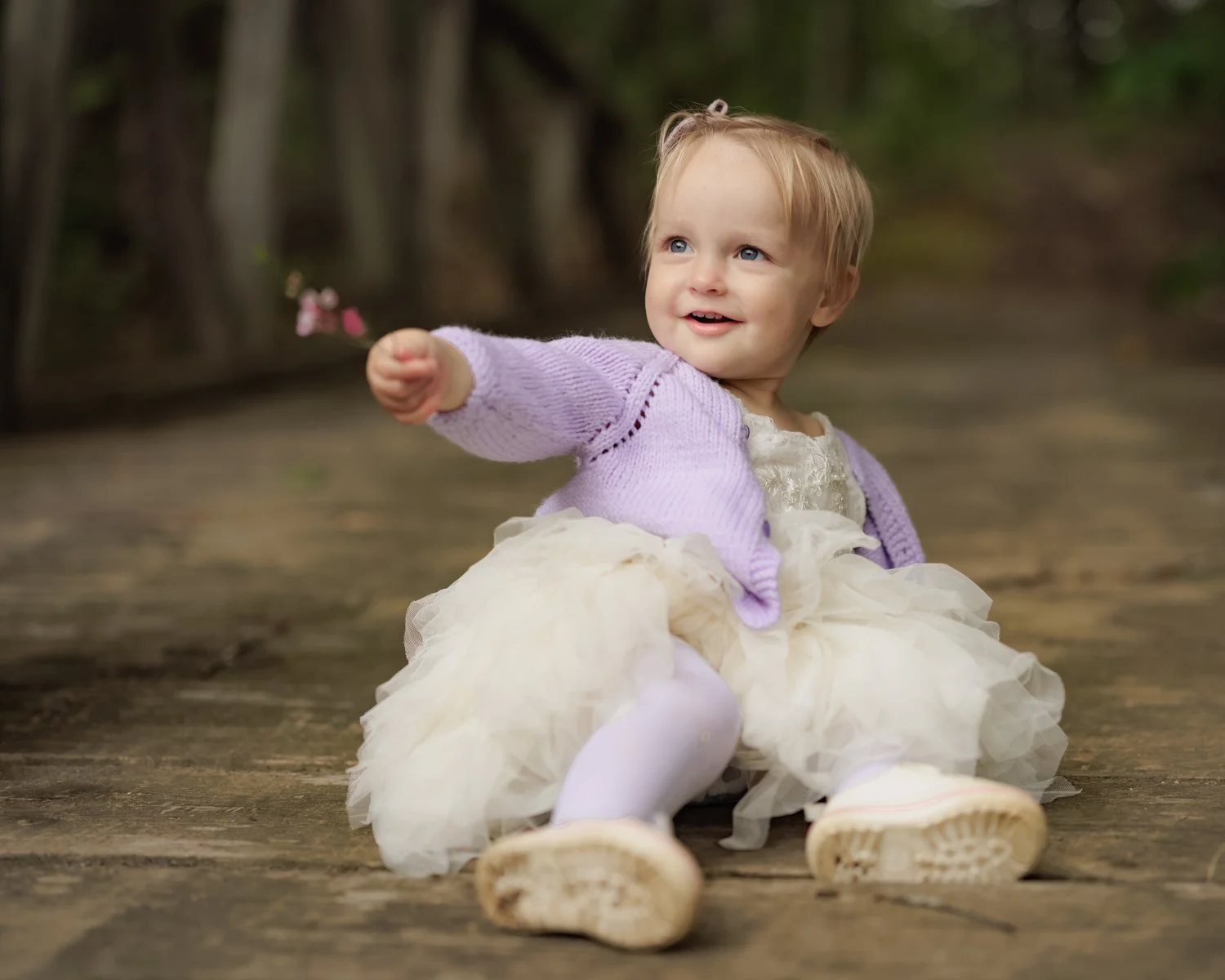 A young girl sitting on a wooden path outdoors, smiling, wearing a cream-colored dress, a purple cardigan, white tights, and shoes, with trees in the background.