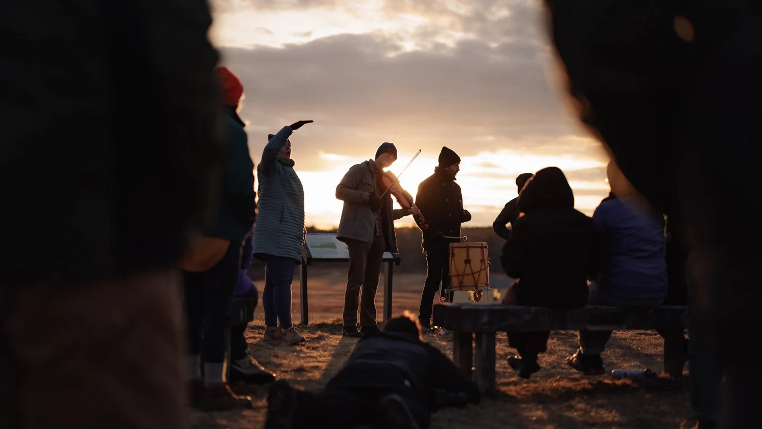 People gathered outdoors at sunset, some standing and others sitting, with musicians playing instruments, including a violin and drums, during an outdoor gathering.