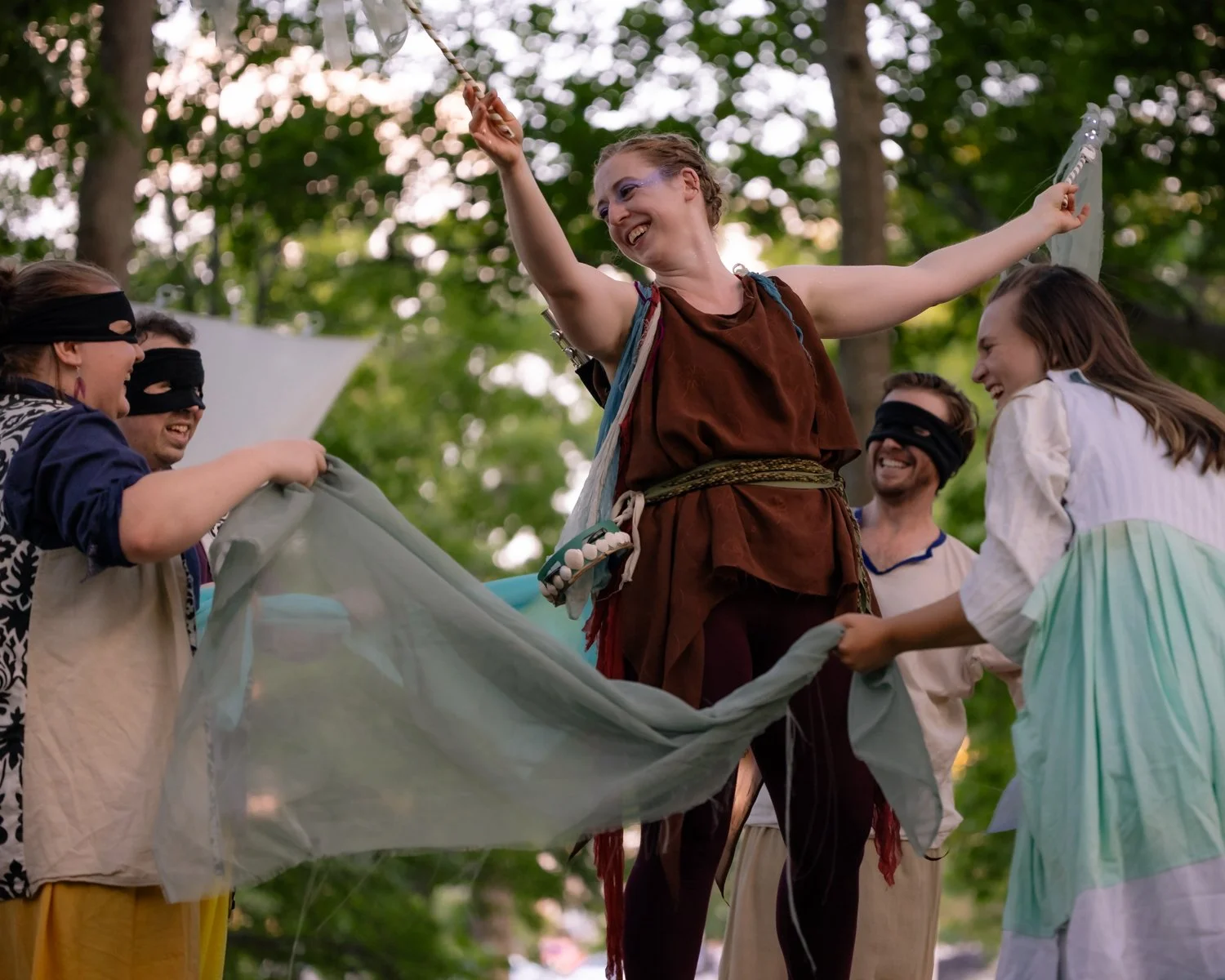 A group of people dressed in medieval or fantasy costumes celebrating outdoors among trees, with some wearing eye masks and holding a fabric.