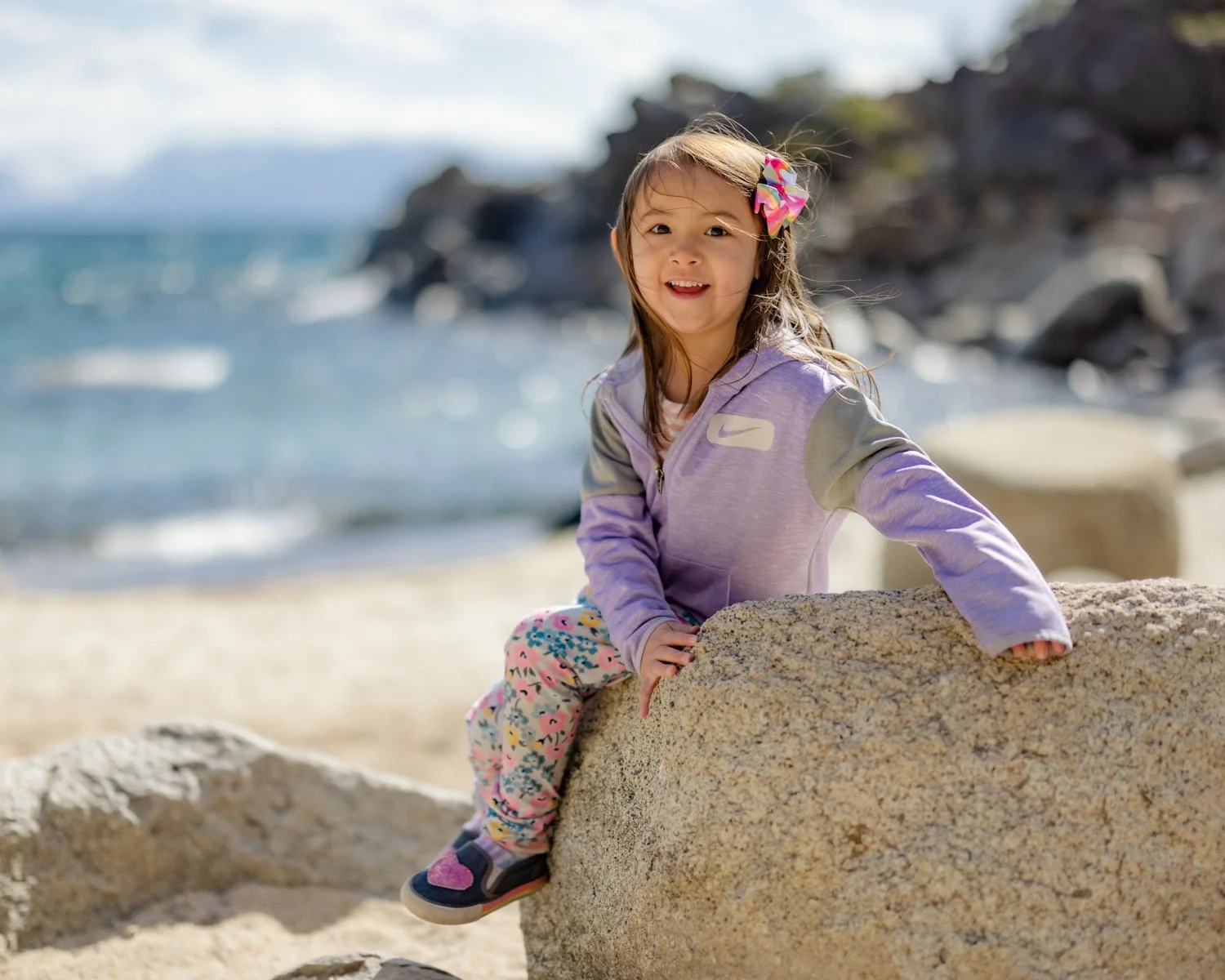 A young girl with long brown hair and a colorful bow, sitting on a large rock at the beach, smiling at the camera with ocean waves and rocky shoreline in the background.