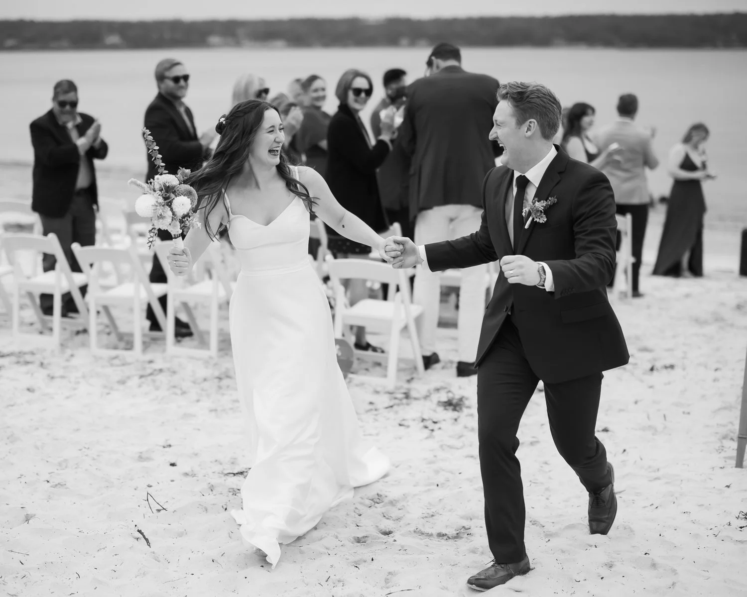A black and white photo of a wedding reception on the beach, showing a smiling bride and groom holding hands and dancing. The bride is wearing a white wedding dress and holding a bouquet, while the groom is dressed in a suit. Guests are in the backgr