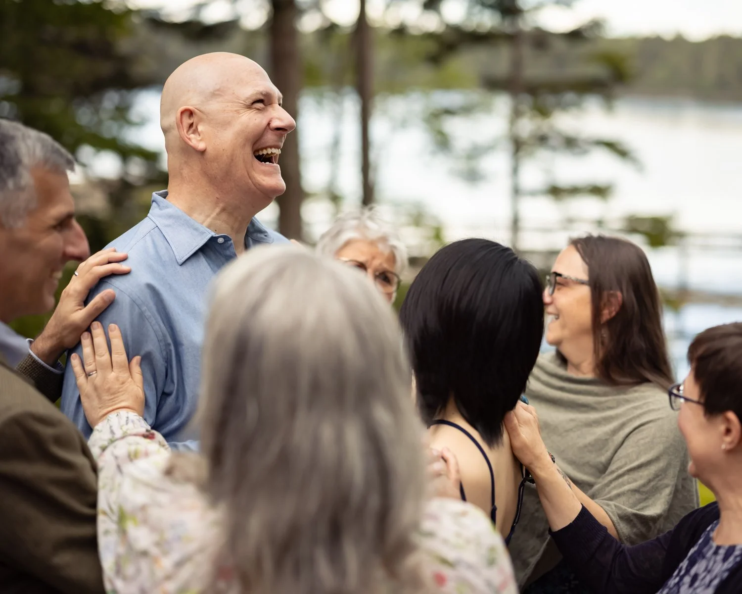A group of people outdoors, smiling and laughing, with trees and water in the background.