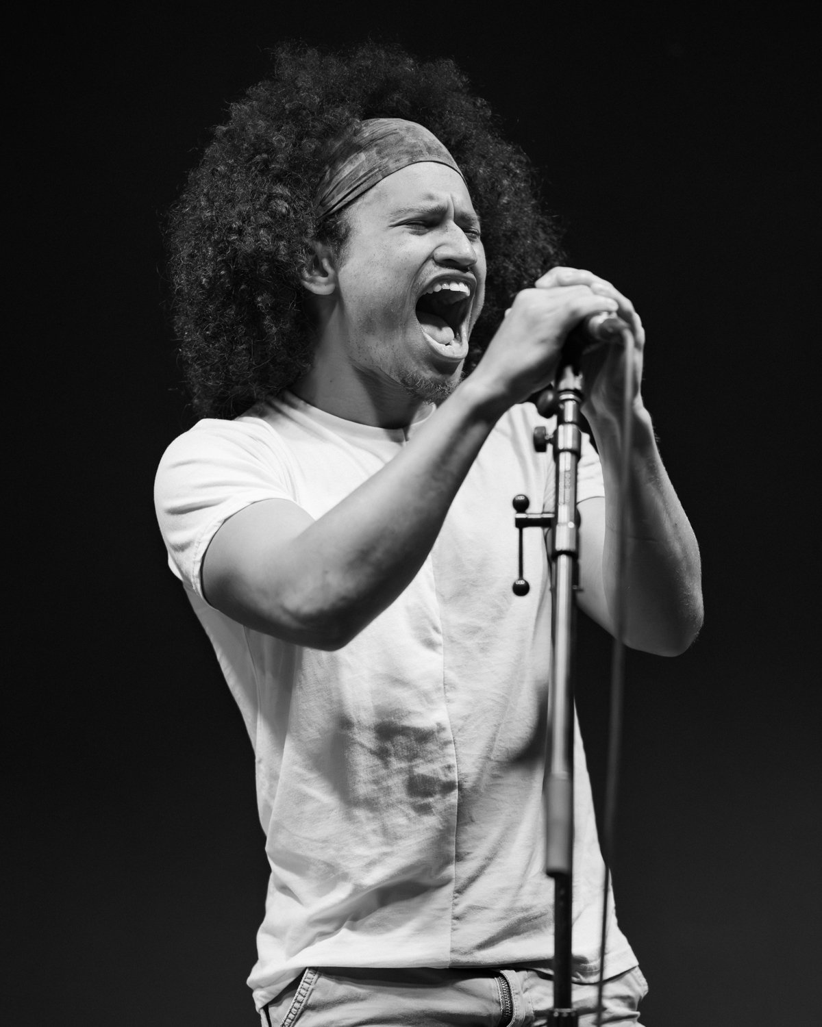 A man with afro hair wearing a headband, singing passionately into a microphone on a dark stage.
