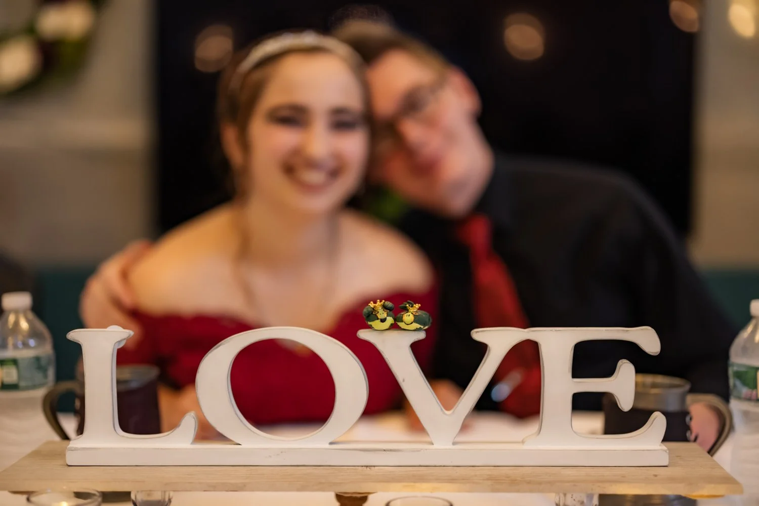 A blurred picture of a smiling woman in a red dress and man in a black shirt and red tie, sitting behind a table with a white decorative 'LOVE' sign on it, topped with small yellow and black bees.