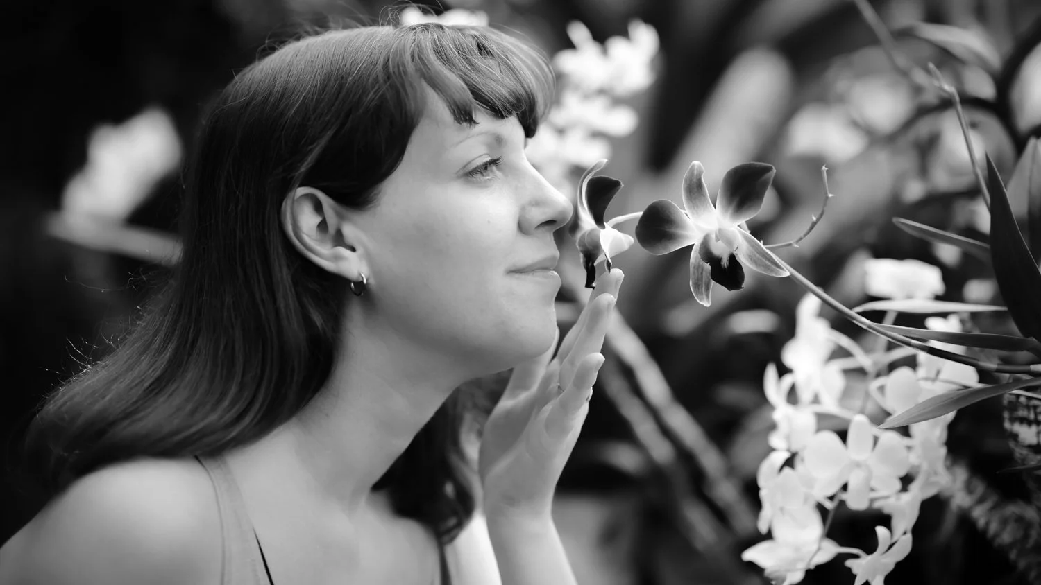 A woman with long dark hair and earrings gently touching an orchid flower in a garden.