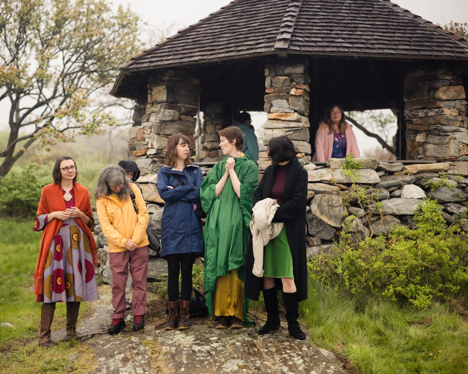 A group of seven women standing and talking outdoors near a stone pavilion, with some wearing rain jackets, in a green grassy area with trees in the background.