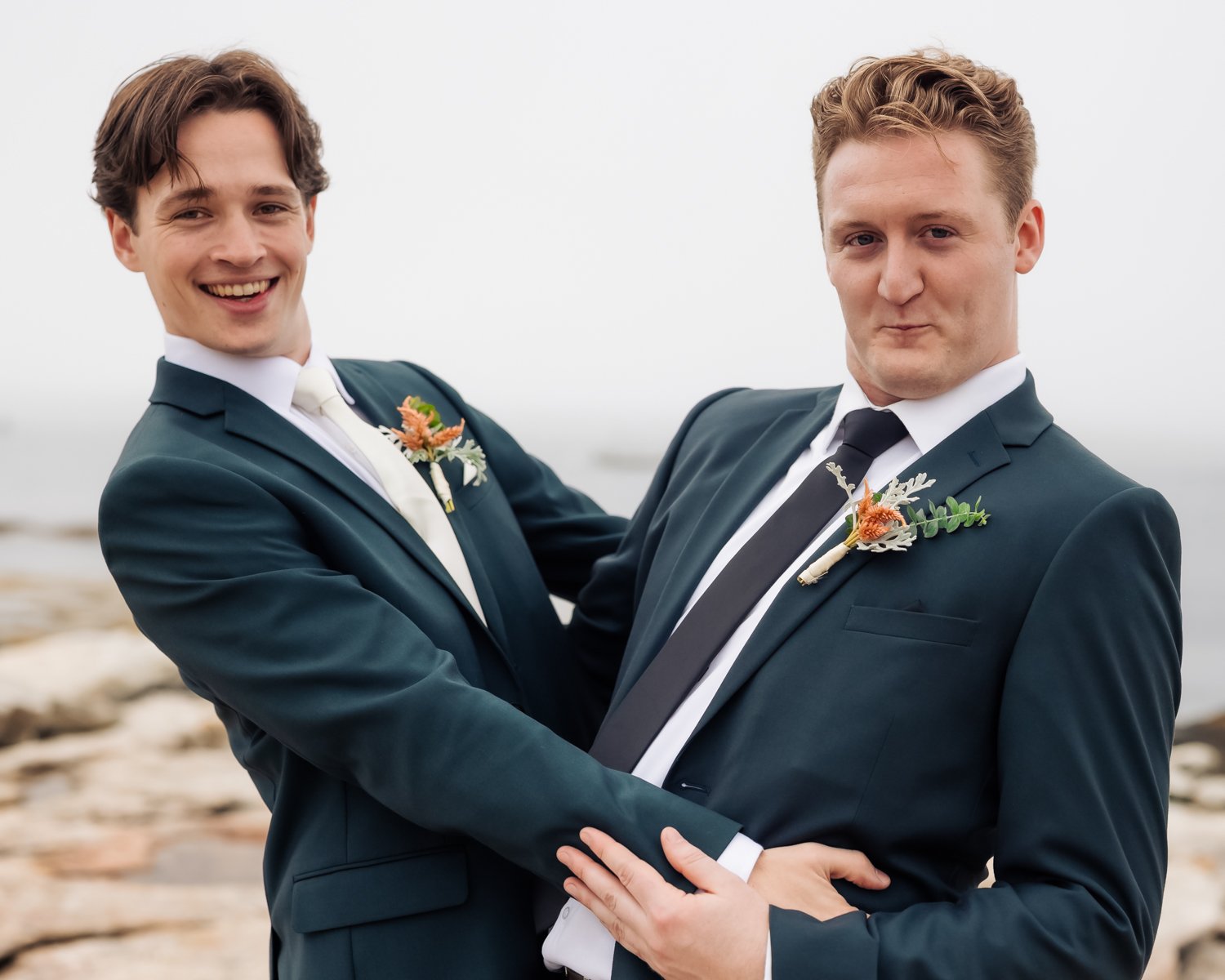 Two men in dark suits, white shirts, and ties holding each other at the beach, both with boutonnières, smiling for the camera.