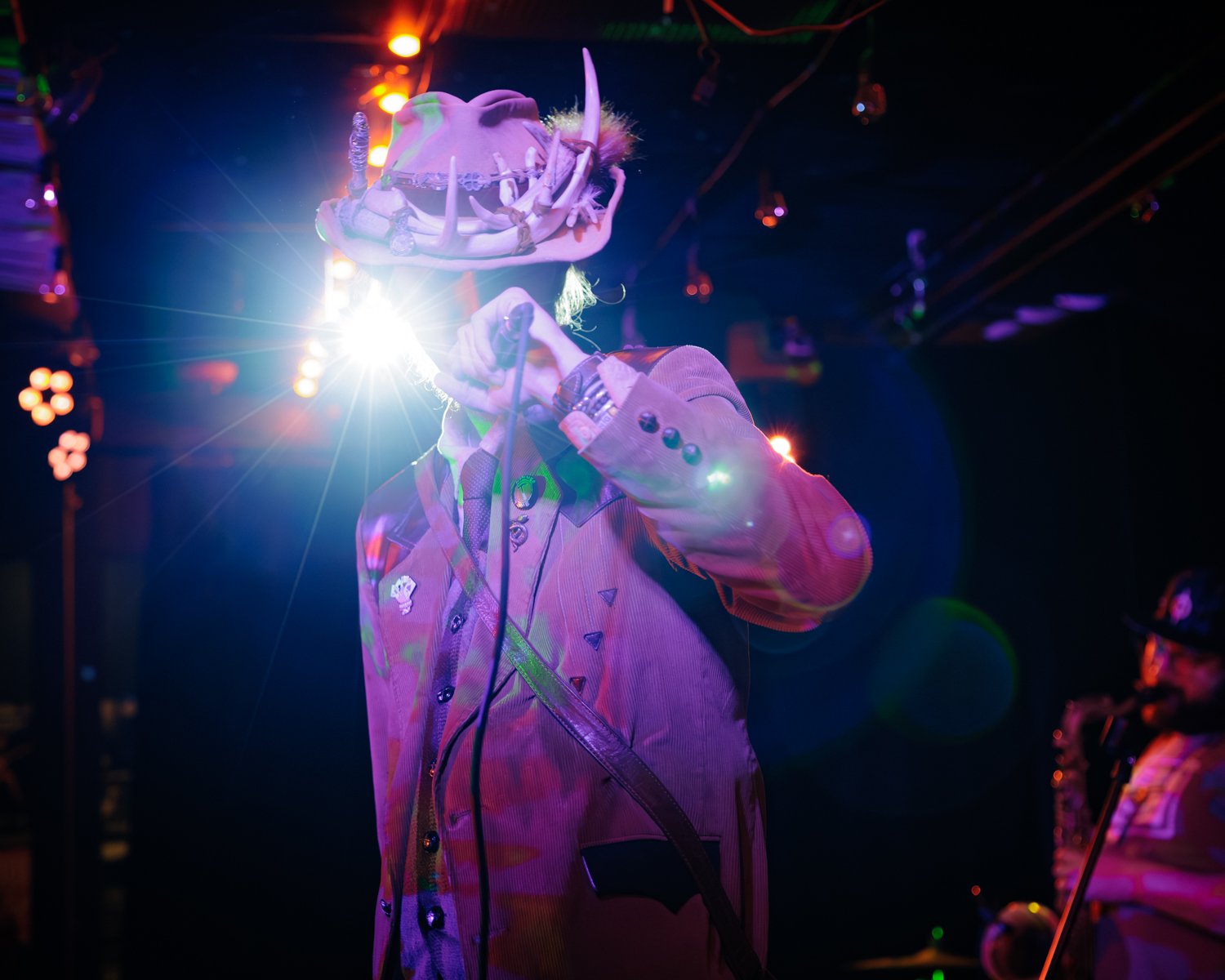Person dressed in a costume with a large, animal skull-like mask and horns, holding a microphone on stage with colorful lighting and lens flares.