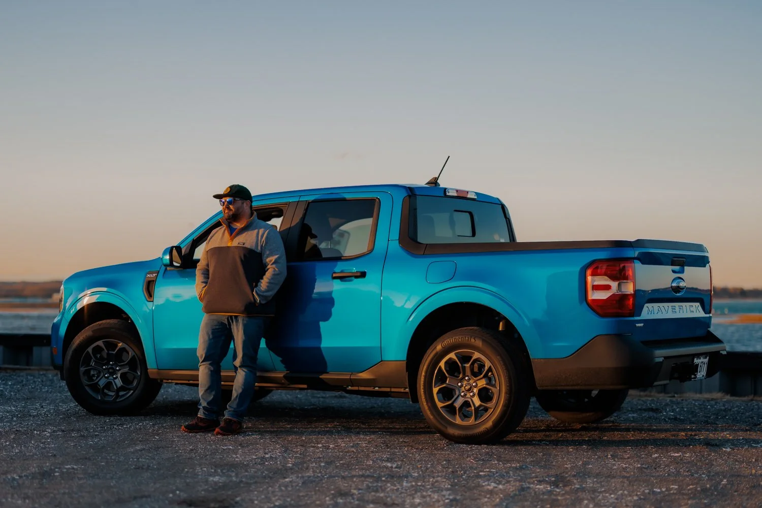 A man in a gray and black jacket, jeans, and sunglasses leaning against a blue Ford Maverick pickup truck on a gravel surface near a body of water during sunset.