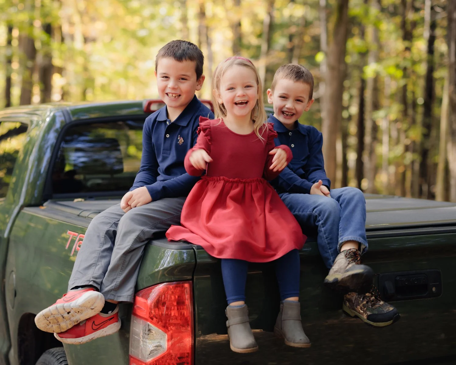 Three children sitting on the back of a dark green pickup truck in a wooded area, smiling and enjoying their time outdoors. The children are dressed casually, with two boys wearing navy shirts and a girl in a red dress.