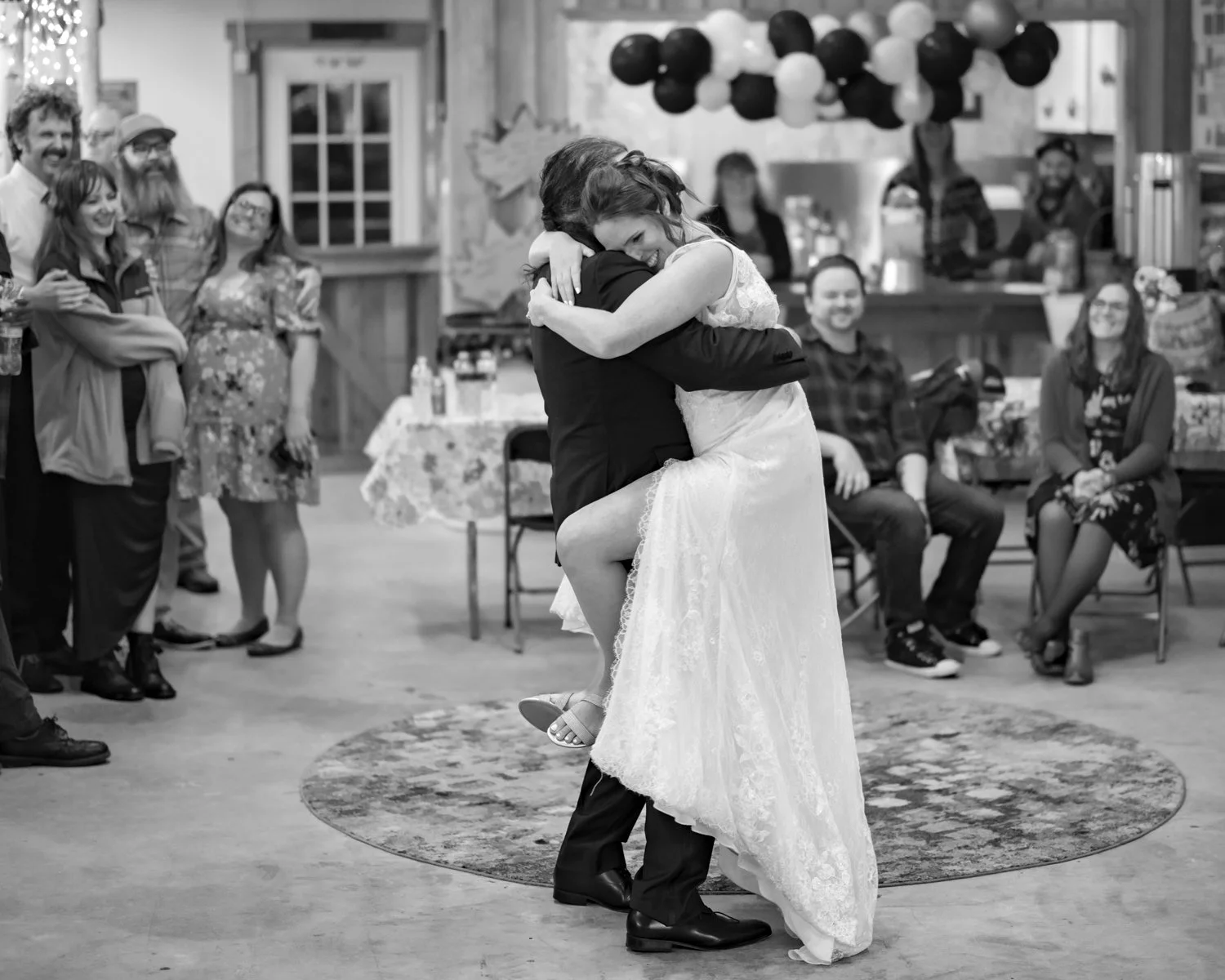 A bride and groom are dancing closely and hugging during their wedding reception. The crowd is watching happily in the background, with some guests seated and others standing.