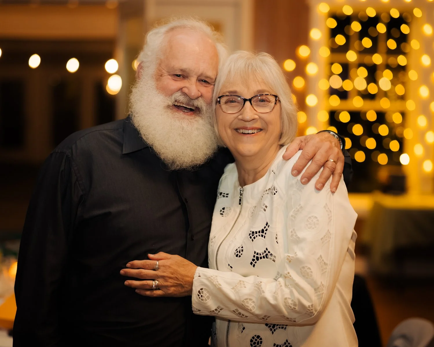 An elderly man and woman smiling and hugging at a warmly lit indoor event with bokeh lights in the background.