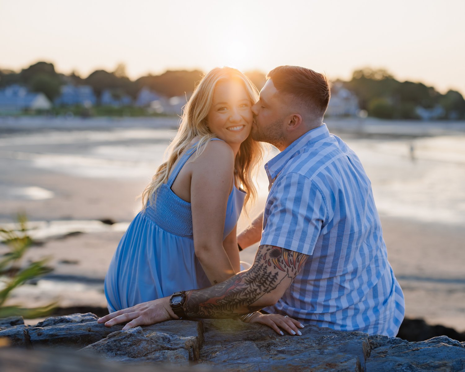 A couple sitting close together on a rock at the beach during sunset, the man kissing the woman's cheek, both wearing blue clothing, with water and houses in the background.