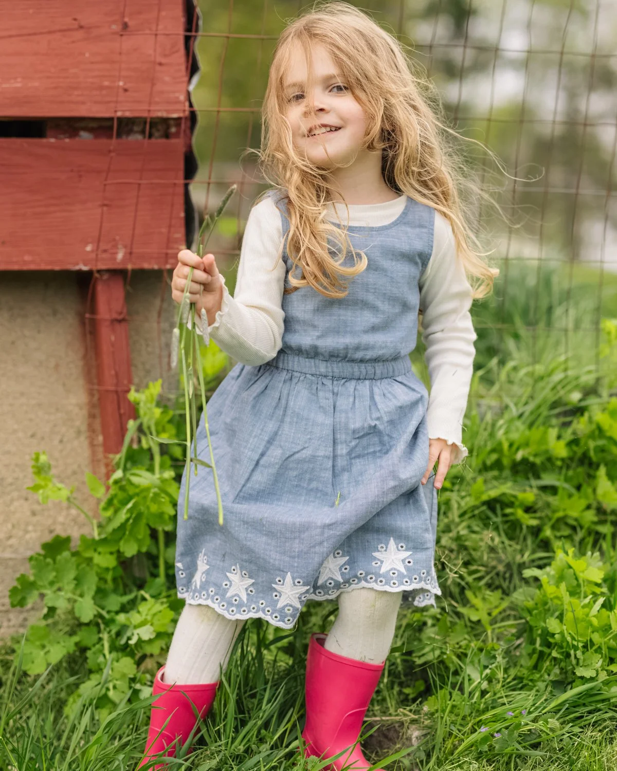 A young girl with long blonde hair wearing a blue dress, white long-sleeve shirt, and pink rain boots standing outdoors near a wooden fence and green plants, holding a small plant in her hand and smiling.