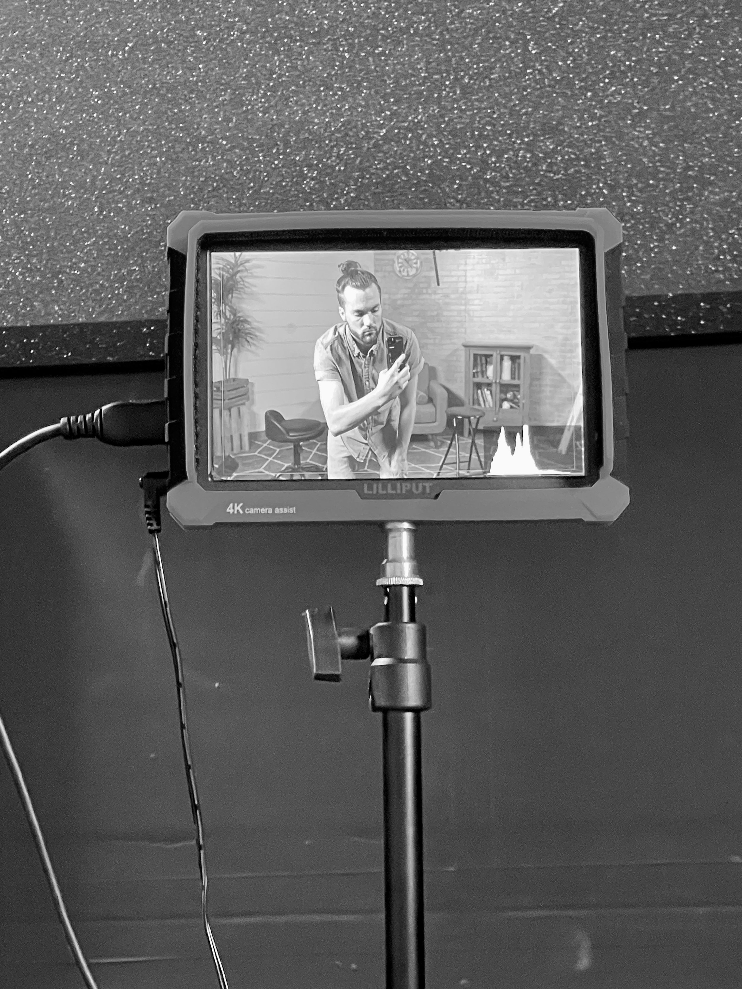 A portable monitor displaying a black and white image of Tristan Mailloux with a top knot hairstyle and a beard, holding a smartphone, sitting in a modern video studio with a plant, bookshelf, and wall clock.