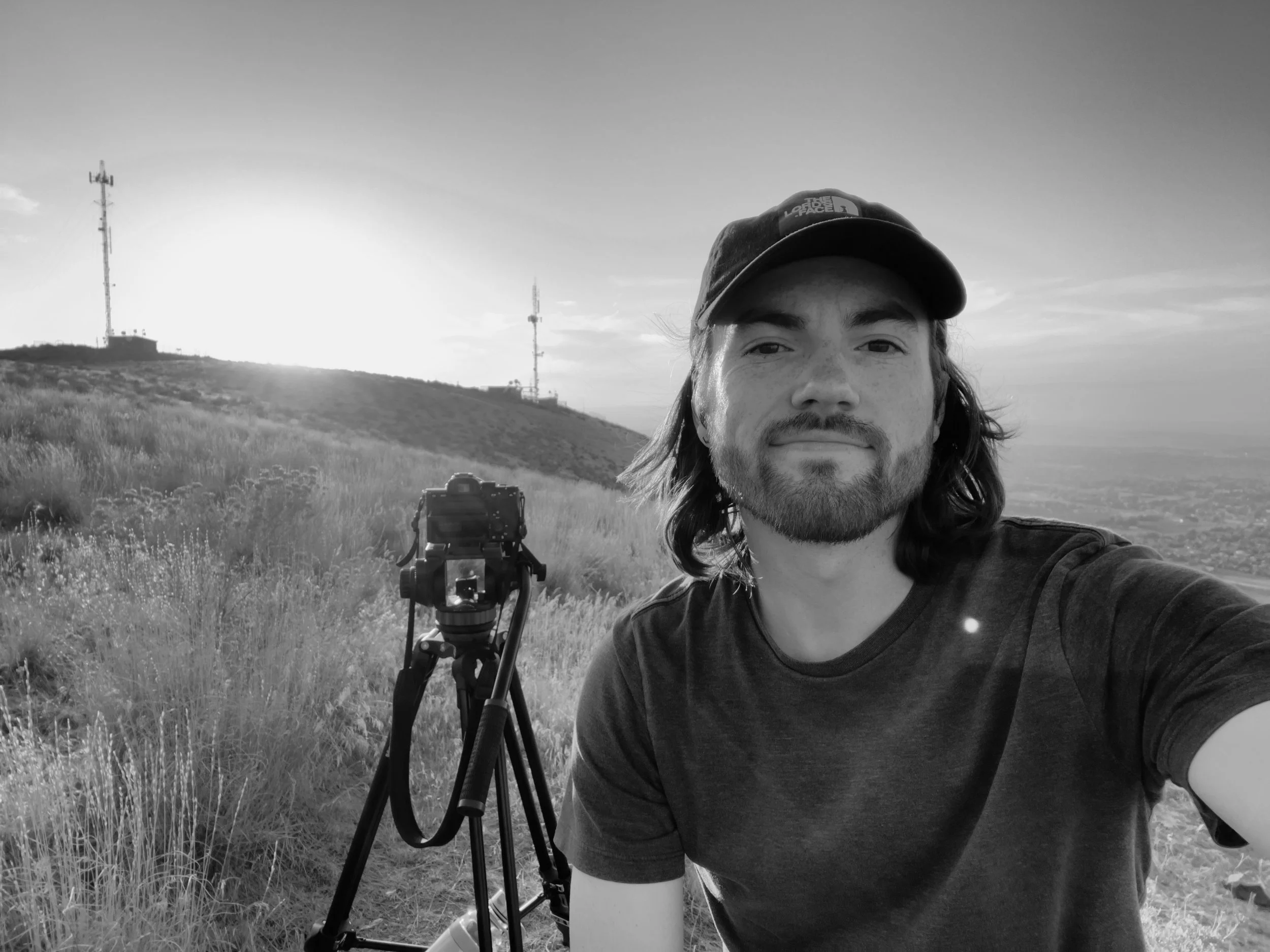 Tristan Mailloux with long hair and a beard taking a selfie outdoors with a camera on a tripod, grassy hillside in the background, and two communication towers on the hilltop, during sunset or sunrise in black and white.