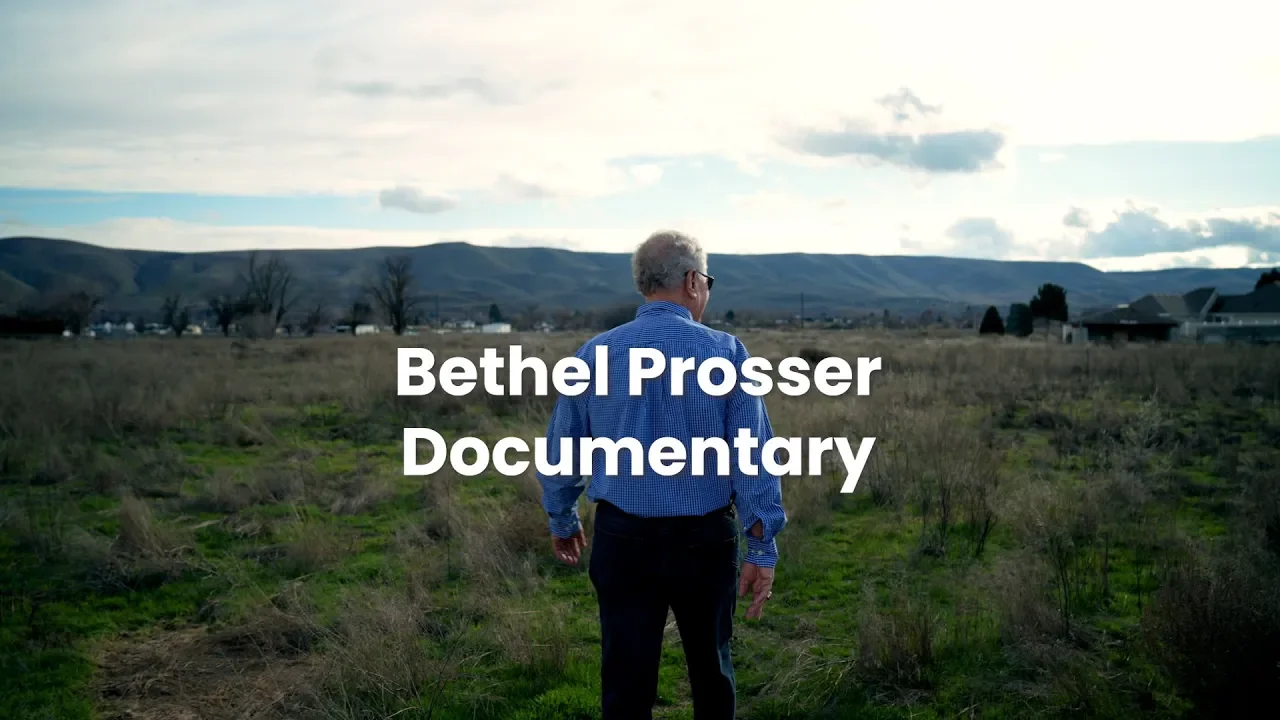 A man with gray hair and glasses walking through an open grassy field, with mountains and a cloudy sky in the background, overlaid with the text 'Bethel Prosser Documentary'.
