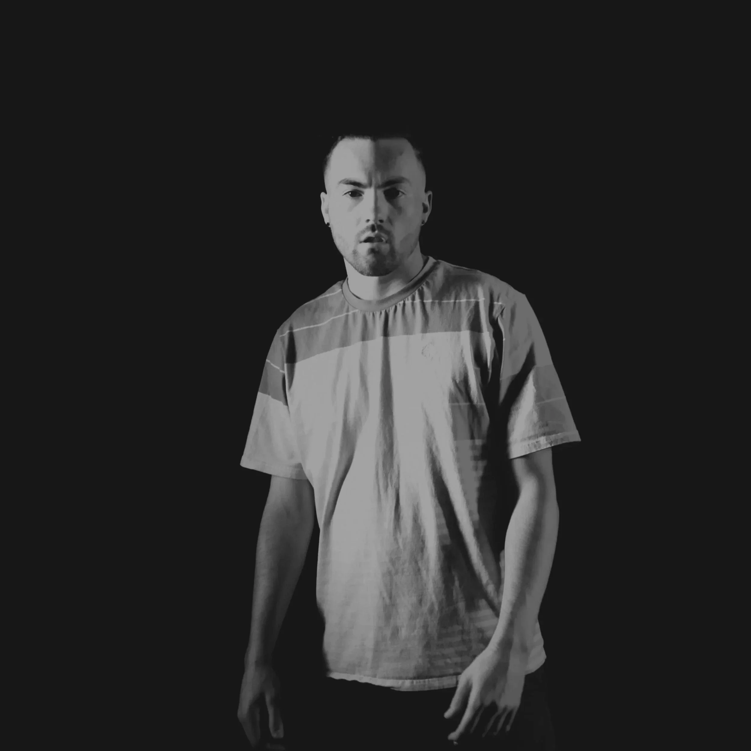 A black and white photo of Tristan Mailloux with short dark hair and a beard, wearing a striped t-shirt, looking at the camera with a serious expression, against a dark background.