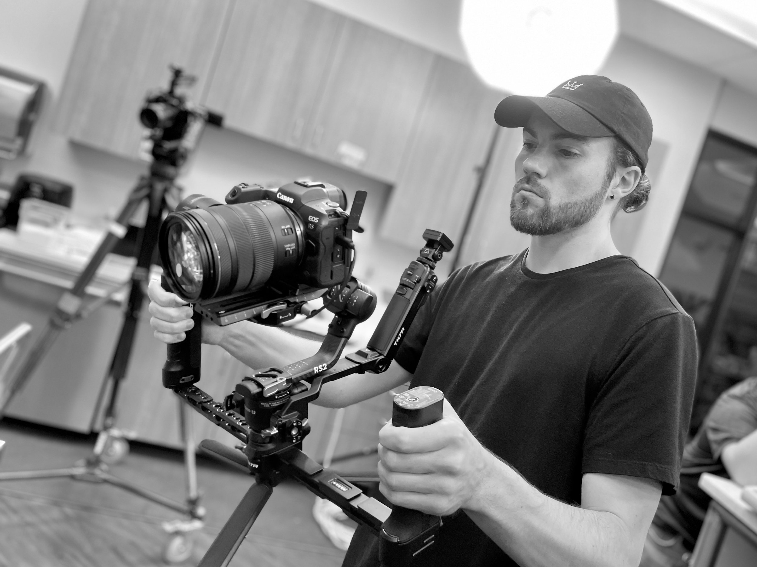 Tristan Mailloux wearing a cap operating a camera stabilizer rig for filming, with background featuring a wooden paneled wall and some equipment.