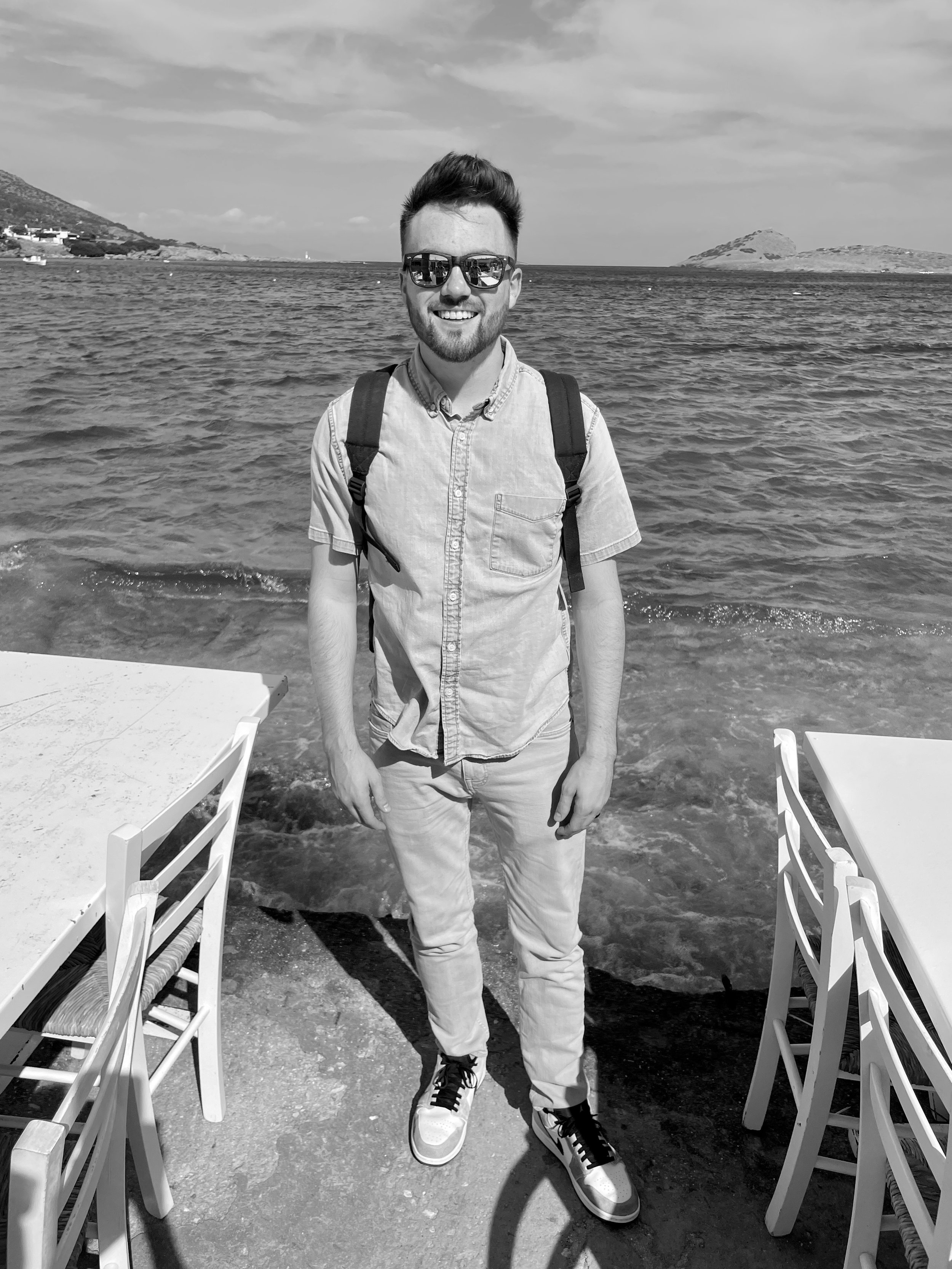 Tristan Mailloux with sunglasses and a backpack standing on a beach, with water and hills in the background.
