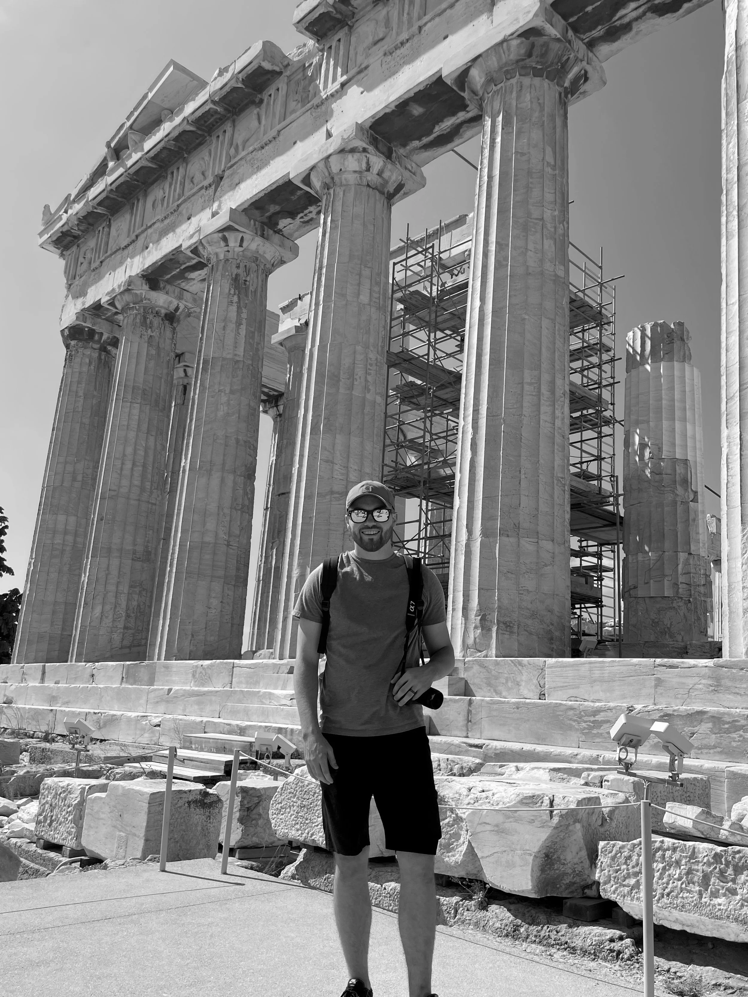 Tristan Mailloux wearing a baseball cap, glasses, a T-shirt, shorts, and a backpack standing in front of ancient Greek ruins, including tall marble columns and scaffolding, at an outdoor archaeological site.