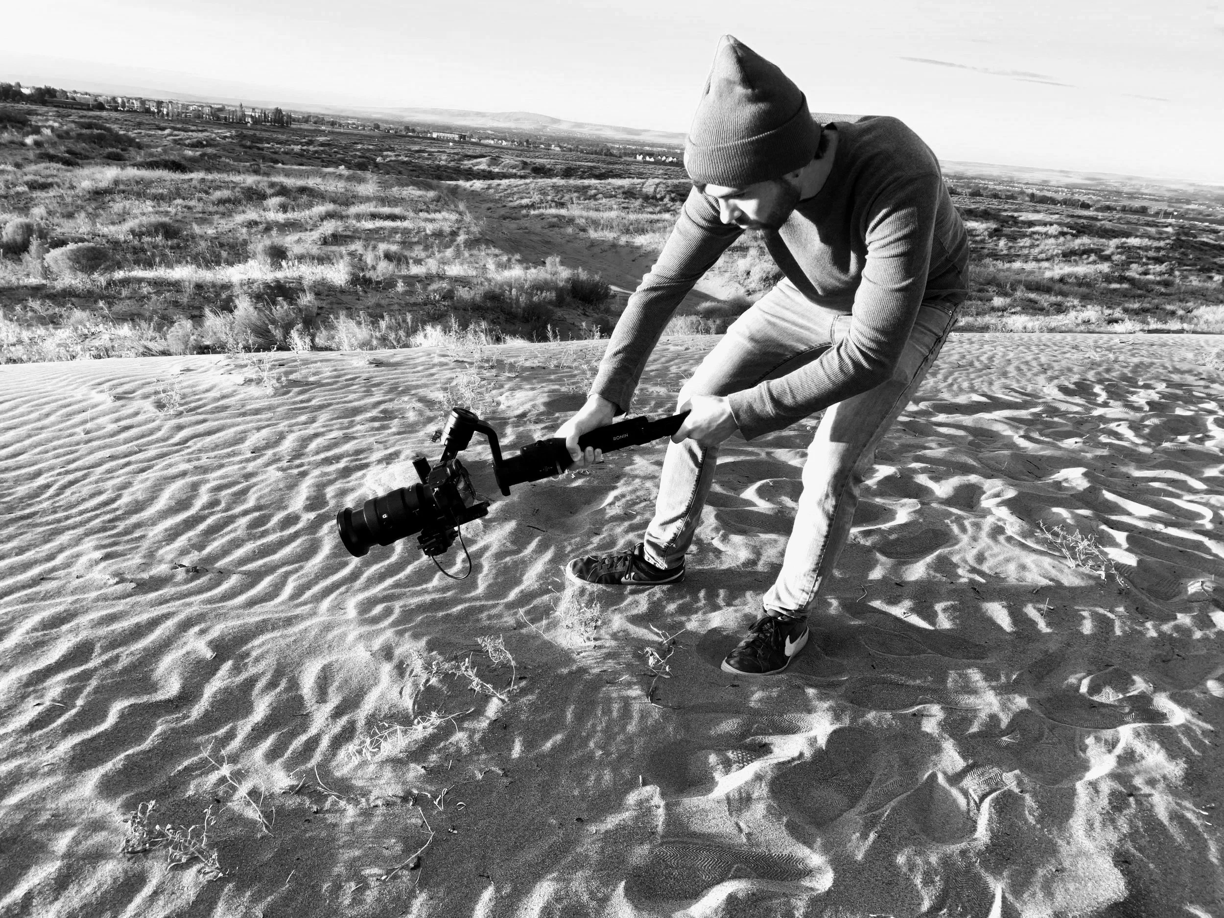 Tristan Mailloux in a beanie and casual clothing bends over on sandy terrain, operating a camera stabilizer with a camera mounted on it, with a landscape of open Pasco land and distant buildings in the background.