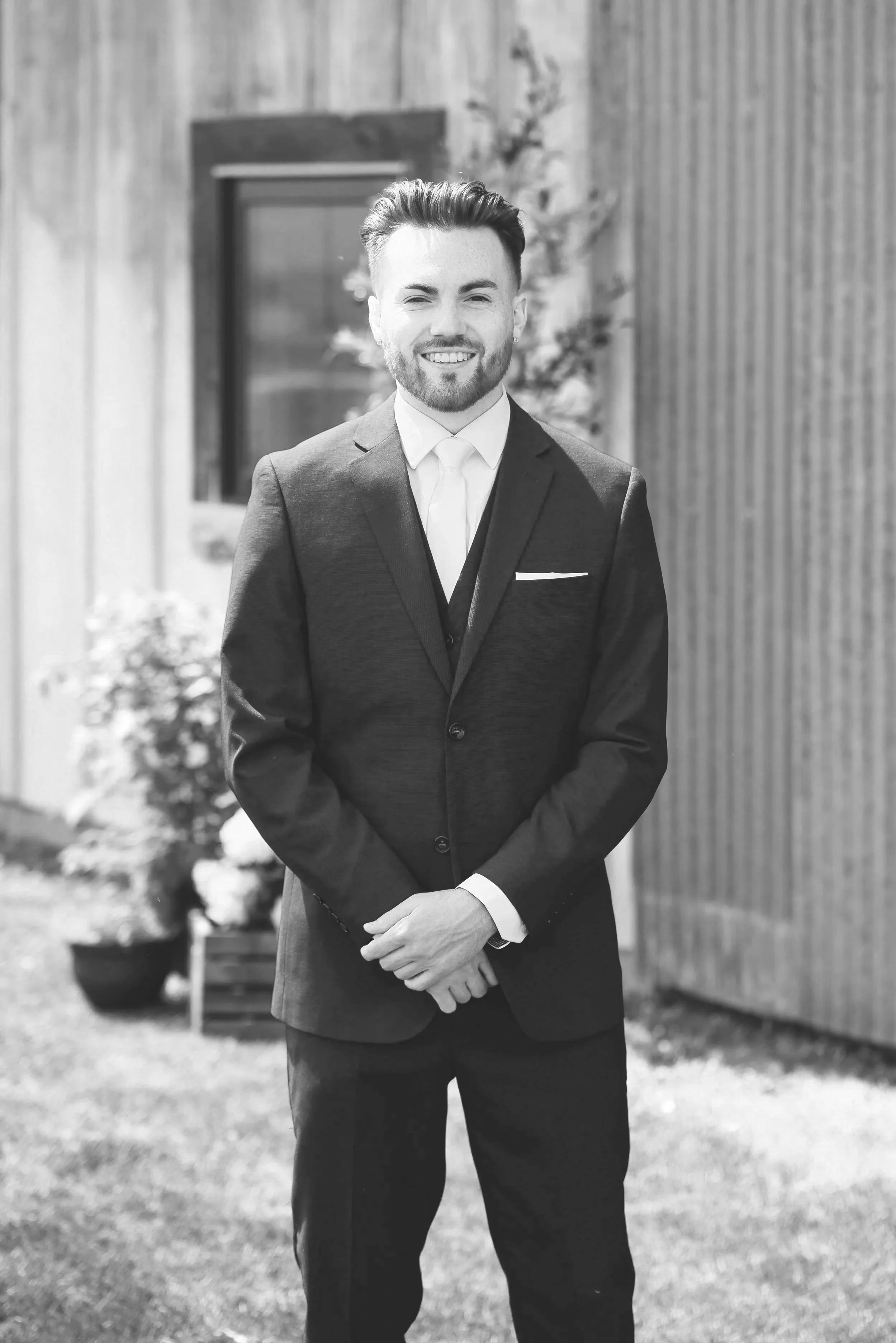Black and white photo of Tristan Mailloux smiling in a suit standing outdoors with a wooden building and potted plant in the background.